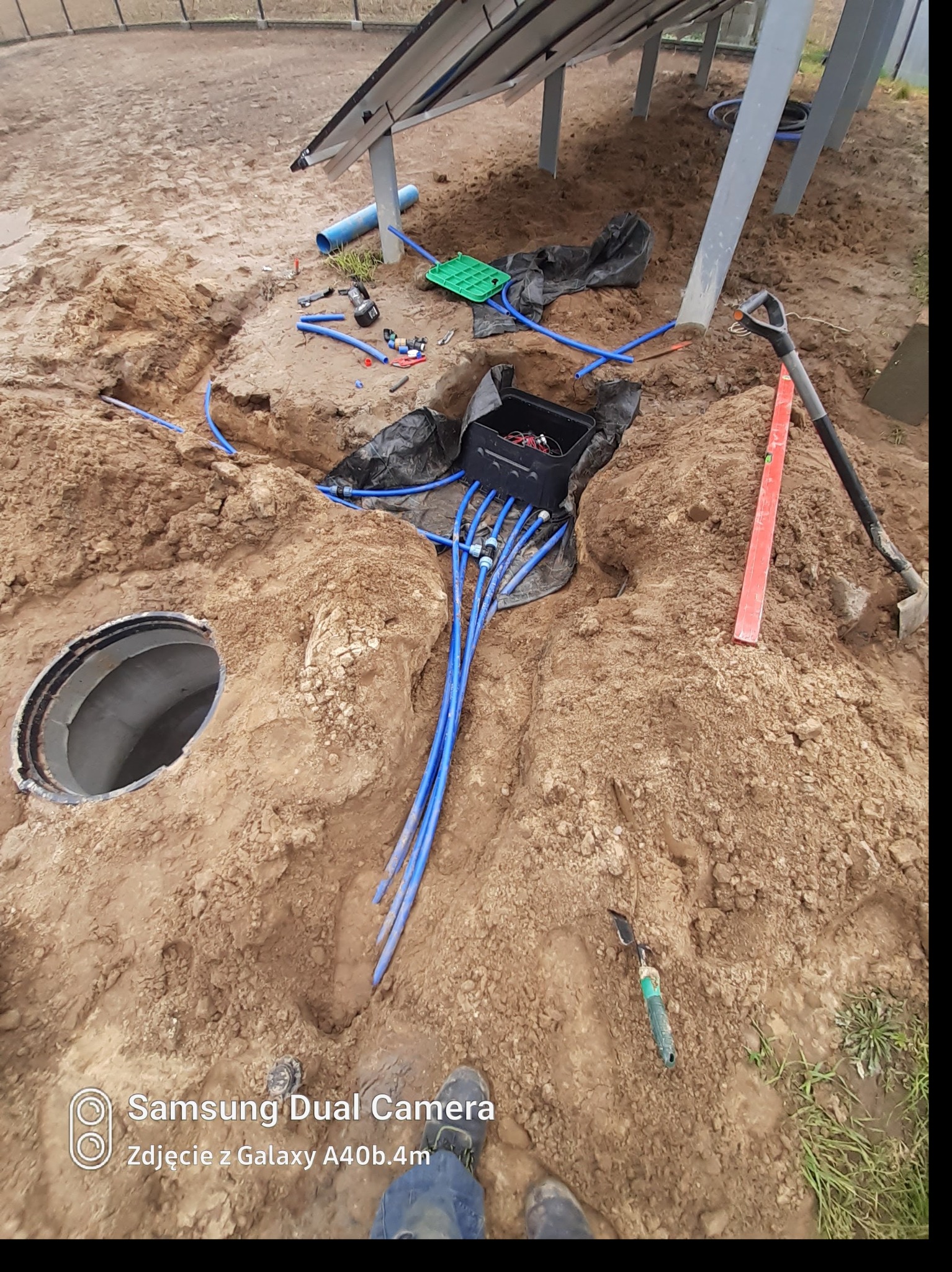 Wykop z widocznymi niebieskimi rurami, czarną skrzynką i narzędziami, prawdopodobnie w trakcie instalacji podziemnego systemu nawadniania ogrodu.