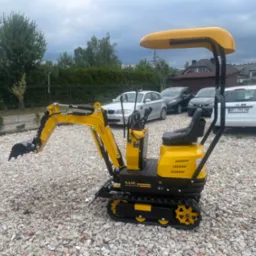 Żółto-czarna mini koparka NANO na żwirowym podłożu, z podniesionym ramieniem koparki. W tle zaparkowane samochody osobowe i zabudowa mieszkalna.