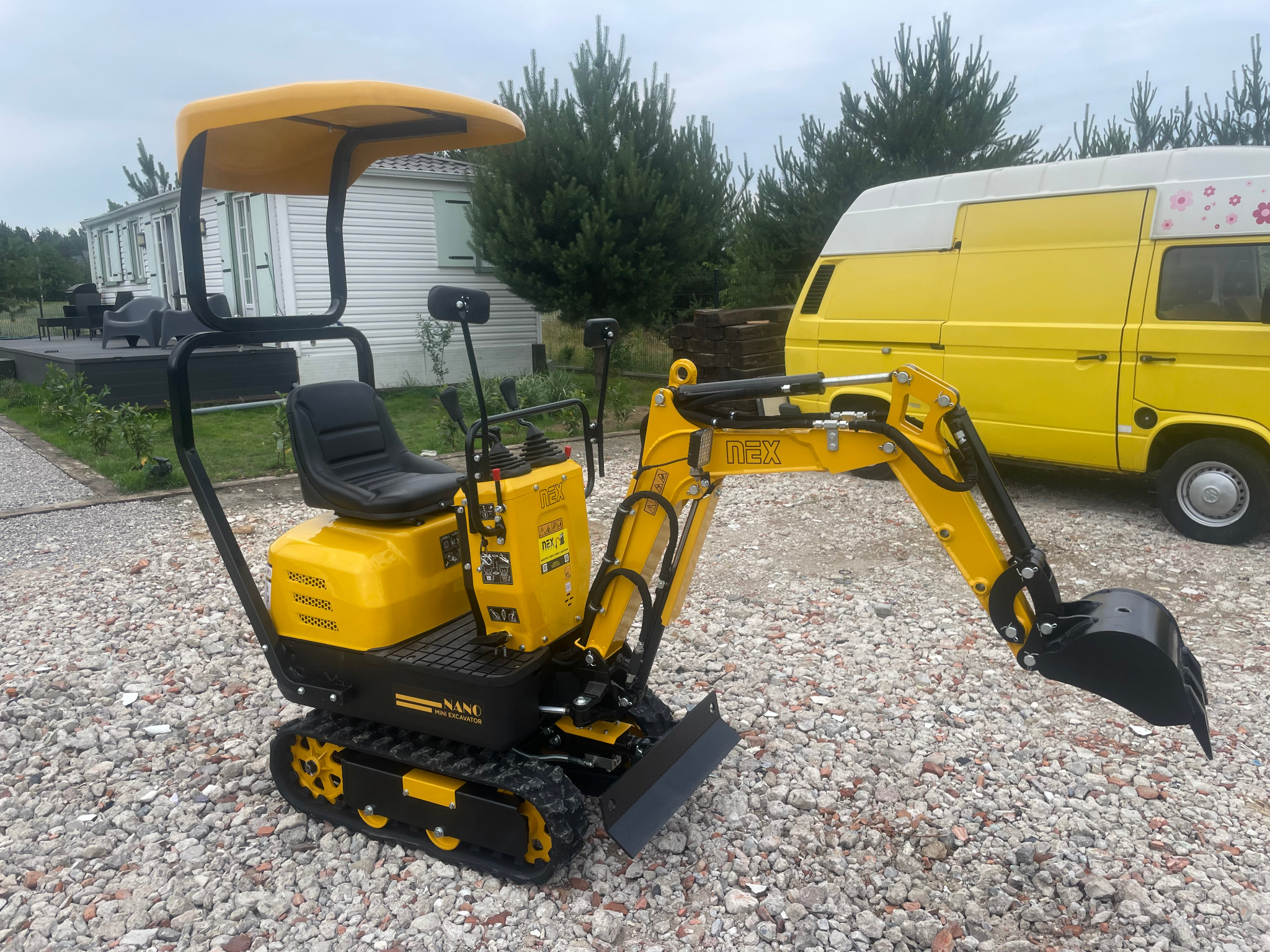 Żółto-czarna mini koparka NANO na żwirowym podłożu, z żółtym busem i domkiem w tle, prezentowana z bliska w naturalnym oświetleniu.