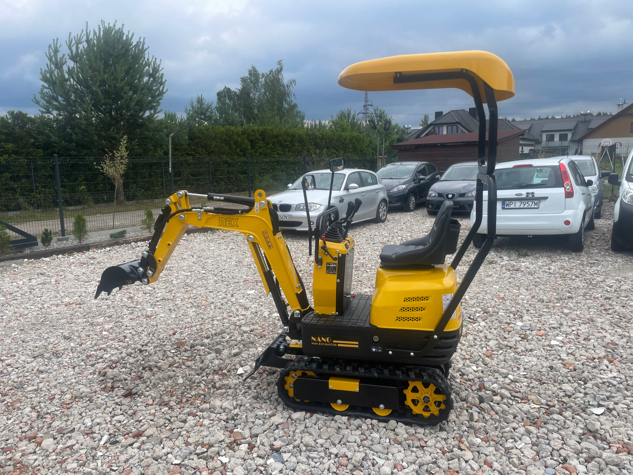 Żółto-czarna mini koparka NANO na żwirowym podłożu, z podniesionym ramieniem koparki. W tle zaparkowane samochody osobowe i zabudowa mieszkalna.