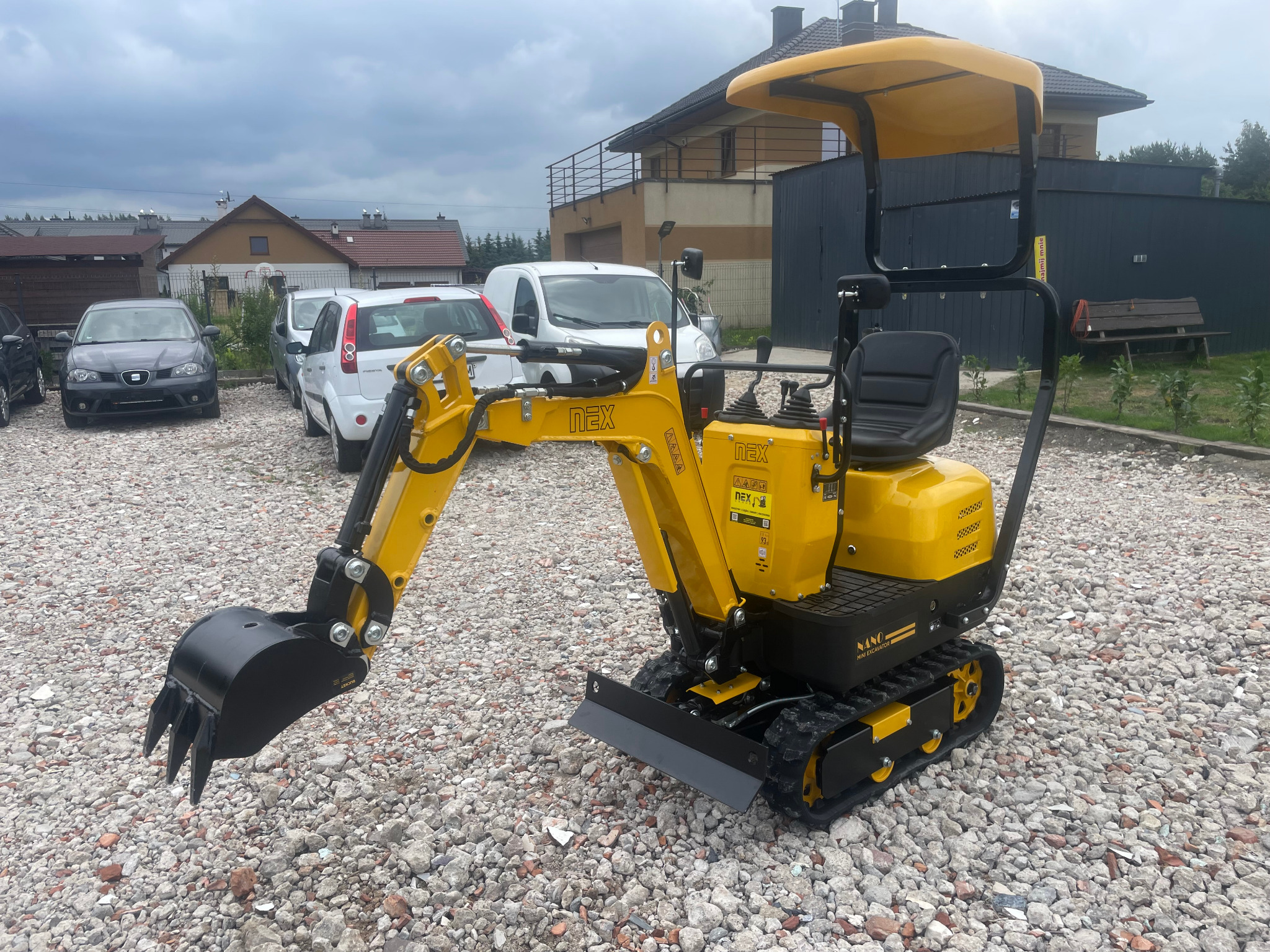 Żółta mini koparka NANO na żwirowym podłożu, z łyżką skierowaną w lewo, na tle budynków i zaparkowanych samochodów w pochmurny dzień.