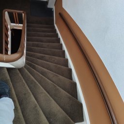 Maltop - Gładzie I Malowanie - Spiralne schody pokryte brązową wykładziną dywanową, z białymi podstopnicami i drewnianą poręczą. Ściany pomalowane na biało i ciepły brąz, widok z góry.