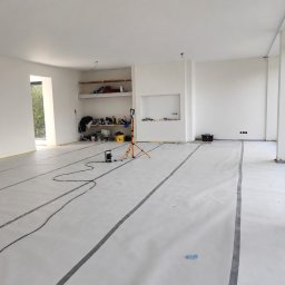 Maltop - Gładzie I Malowanie - Jasne wnętrze po gładziach i malowaniu, z zabezpieczoną szarym filcem podłogą z czarnymi taśmami. Widoczny statyw z lampą, narzędzia i wnęka z półkami.