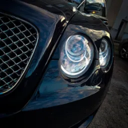 Detal czarnego Bentley'a: reflektor z chromowaną obudową, maska odbijająca niebo i budynek w tle, uchwycony z niskiego kąta.