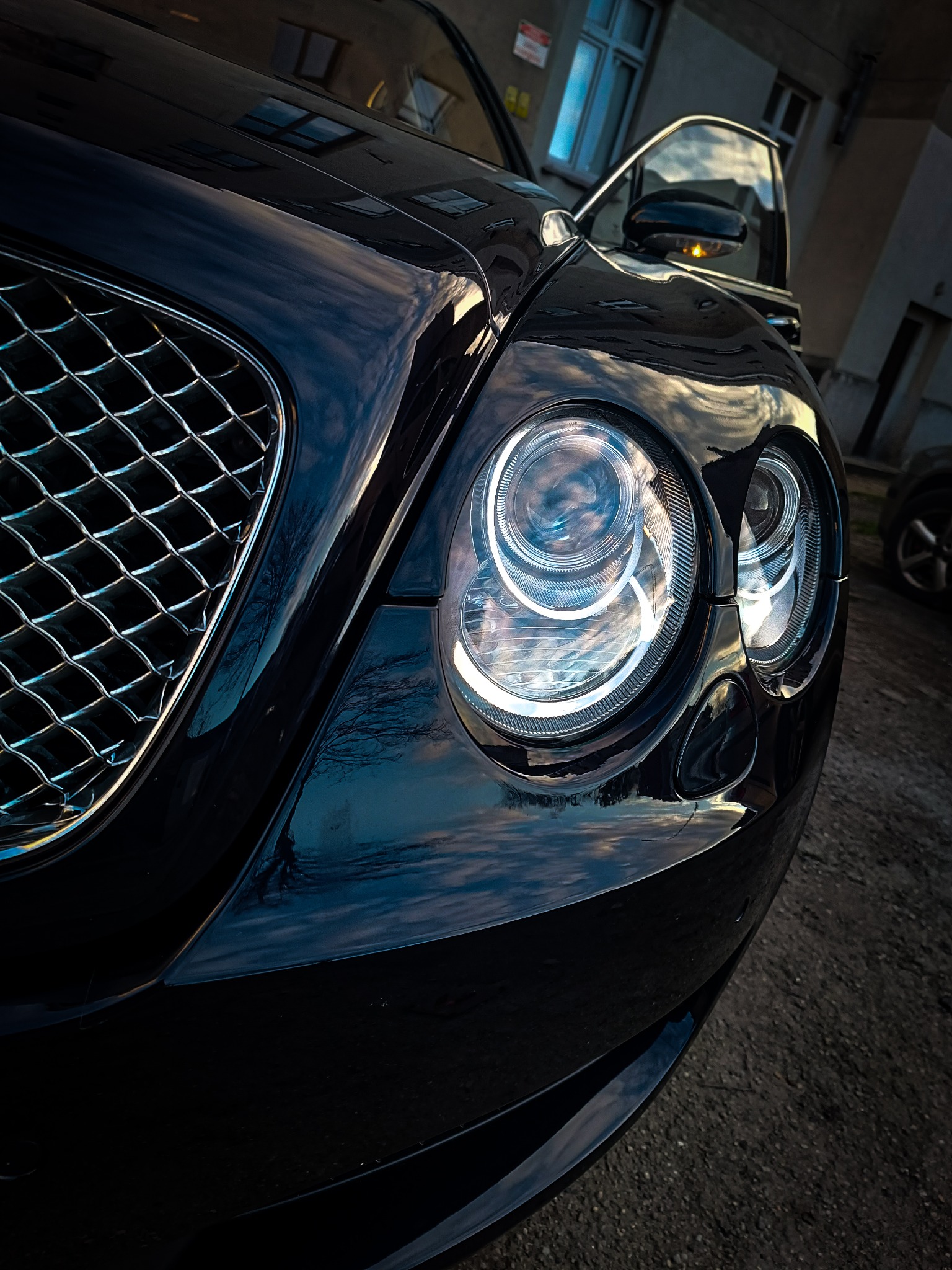 Detal czarnego Bentley'a: reflektor z chromowaną obudową, maska odbijająca niebo i budynek w tle, uchwycony z niskiego kąta.