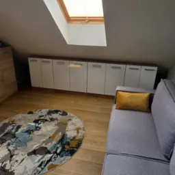 Pok&oacute;j na poddaszu z drewnianą podłogą, szarą sofą i oknem dachowym z żaluzjami. Białe szafki w zabudowie pod skosem. Kolorowy okrągły dywan na podłodze.