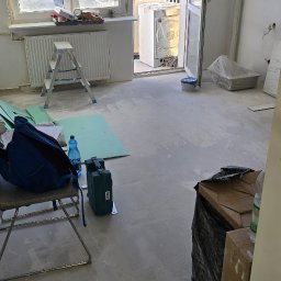 Masters - Wnętrze w trakcie remontu: drabina, narzędzia, materiały podłogowe, otwarte drzwi balkonowe z widokiem na zieleń i budynek. Podłoga przygotowana pod układanie.