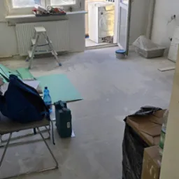 Wnętrze w trakcie remontu: drabina, narzędzia, materiały podłogowe, otwarte drzwi balkonowe z widokiem na zieleń i budynek. Podłoga przygotowana pod układanie.