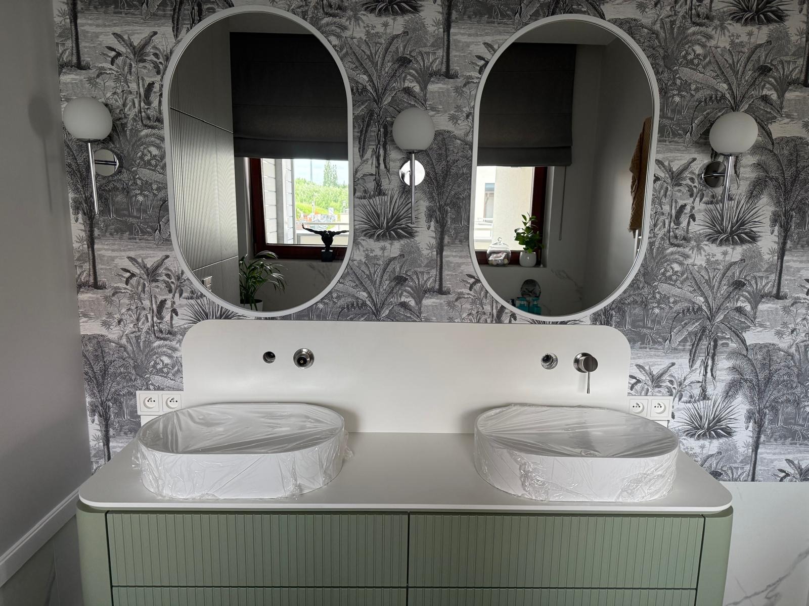 Nowoczesna łazienka z podwójną umywalką i lustrami owalnymi na tle tapety w tropikalne wzory. Umywalki zabezpieczone folią ochronną, zielona szafka pod umywalką.