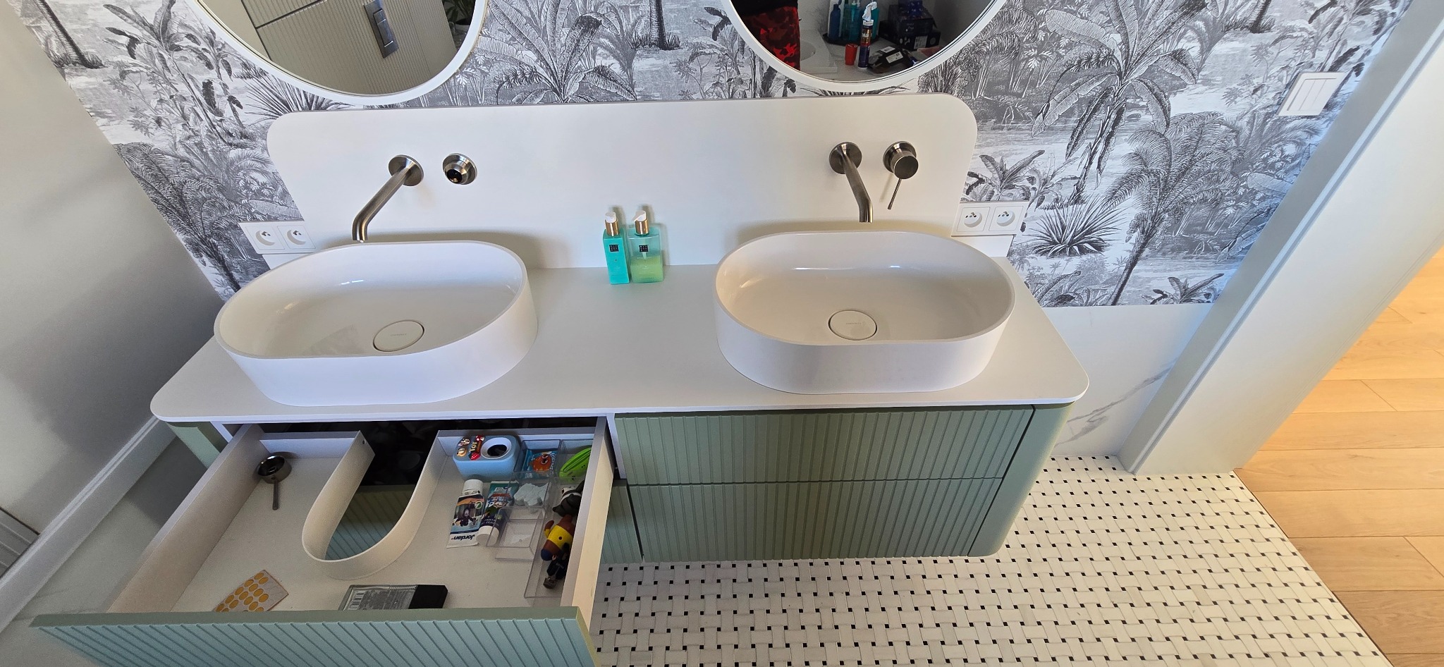 Nowoczesna łazienka z podwójną umywalką nablatową, zieloną szafką z szufladami i wzorzystą tapetą w tropikalnym stylu. Otwarta szuflada z akcesoriami.