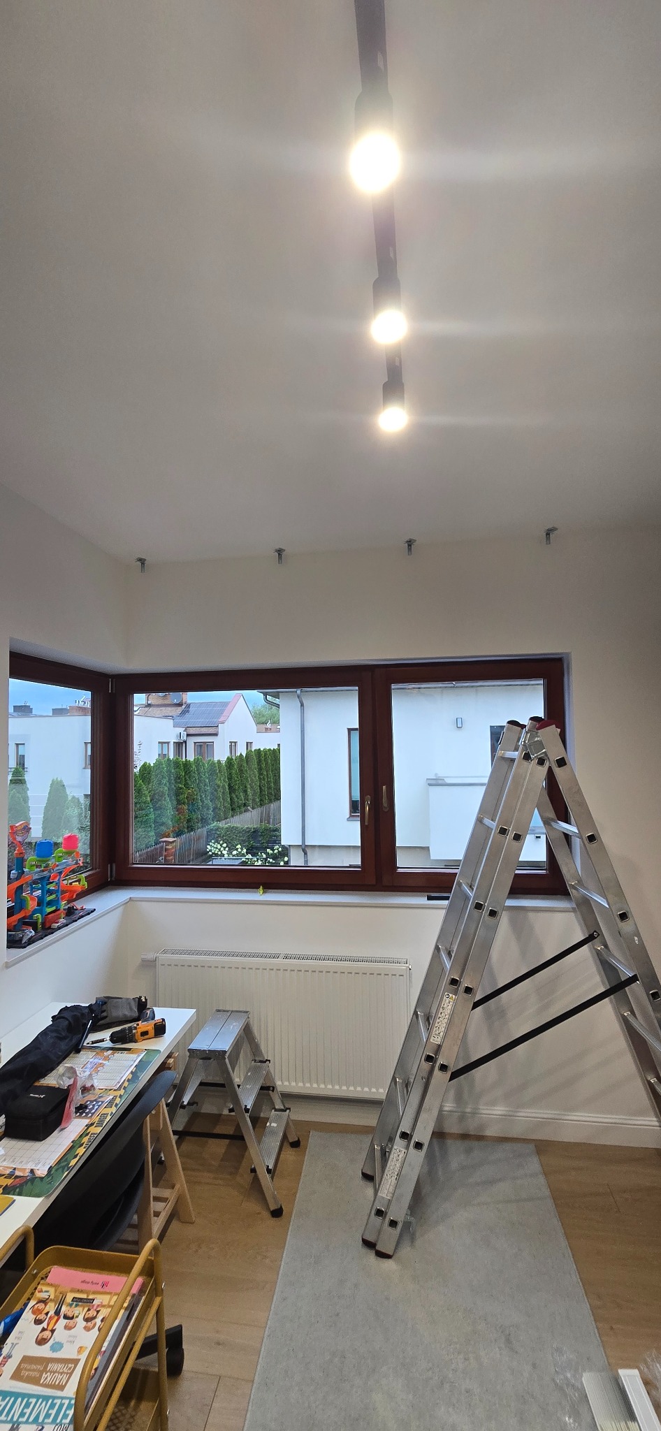 Montaż oświetlenia szynowego w pokoju z oknem. Widoczne narzędzia, drabina i zamontowane uchwyty do szyn na suficie. Praca w toku, widok na zewnątrz.