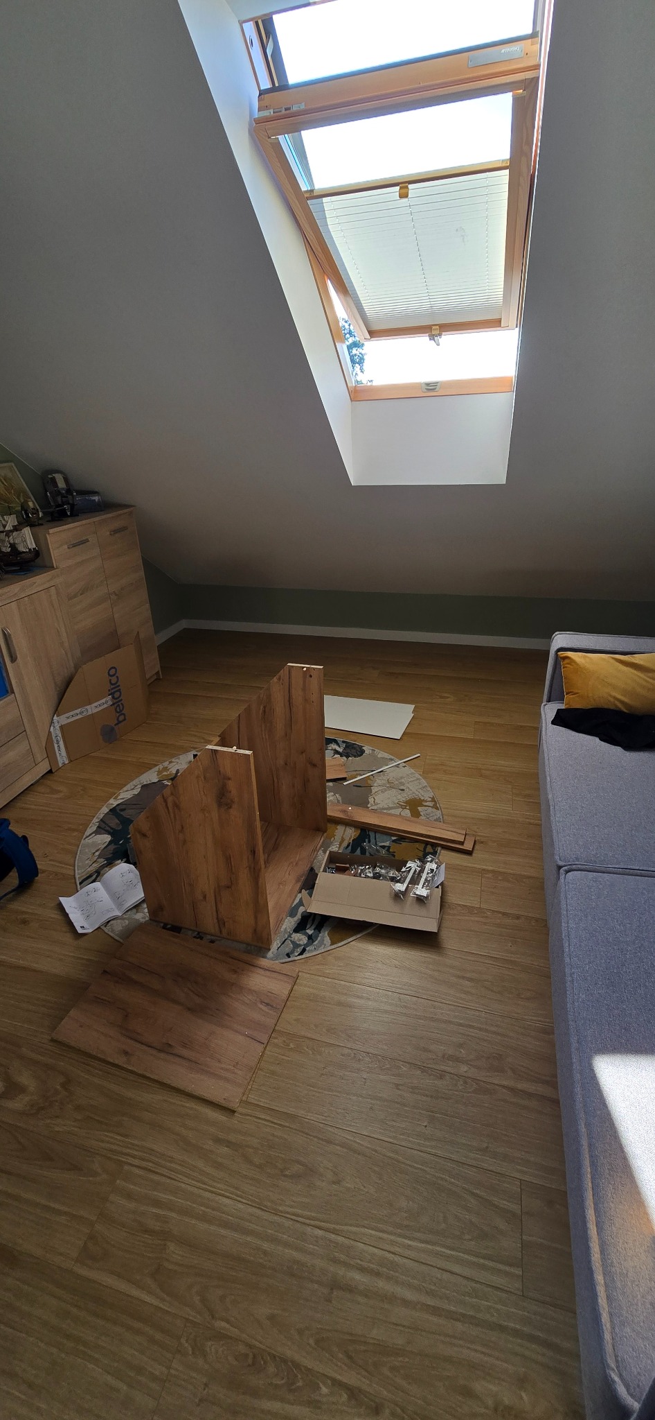 Montaż szafki z drewnopodobnej płyty w pokoju na poddaszu z oknem dachowym. Elementy szafki, instrukcja i akcesoria montażowe rozłożone na podłodze.