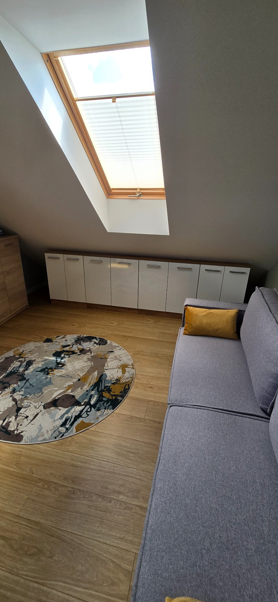 Pokój na poddaszu z drewnianą podłogą, szarą sofą i oknem dachowym z żaluzjami. Białe szafki w zabudowie pod skosem. Kolorowy okrągły dywan na podłodze.