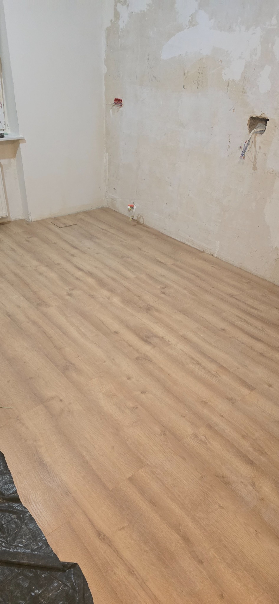 Jasne panele podłogowe w pokoju z surowymi ścianami, widoczne gniazdka elektryczne. Częściowo zakryte folią zabezpieczającą.