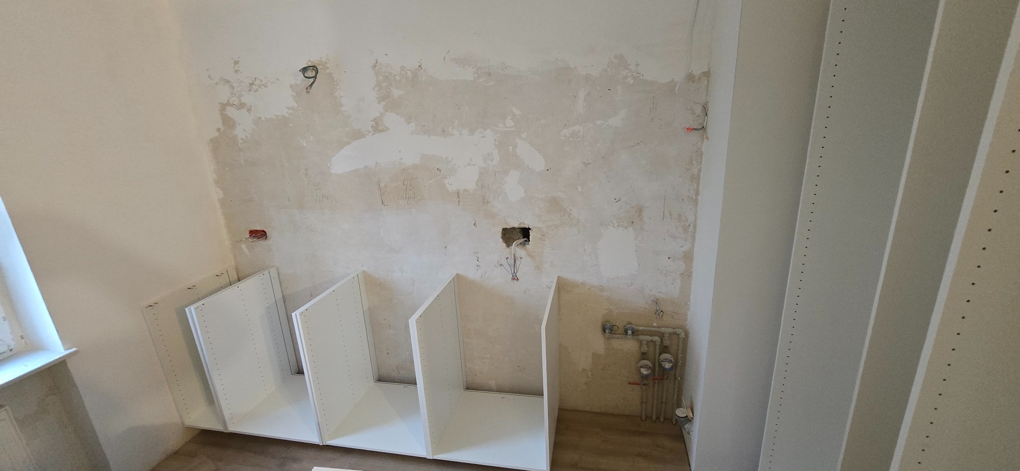 Montaż szafek kuchennych: surowa ściana z instalacjami, częściowo zamontowane białe korpusy szafek, widoczne nawiercenia montażowe.