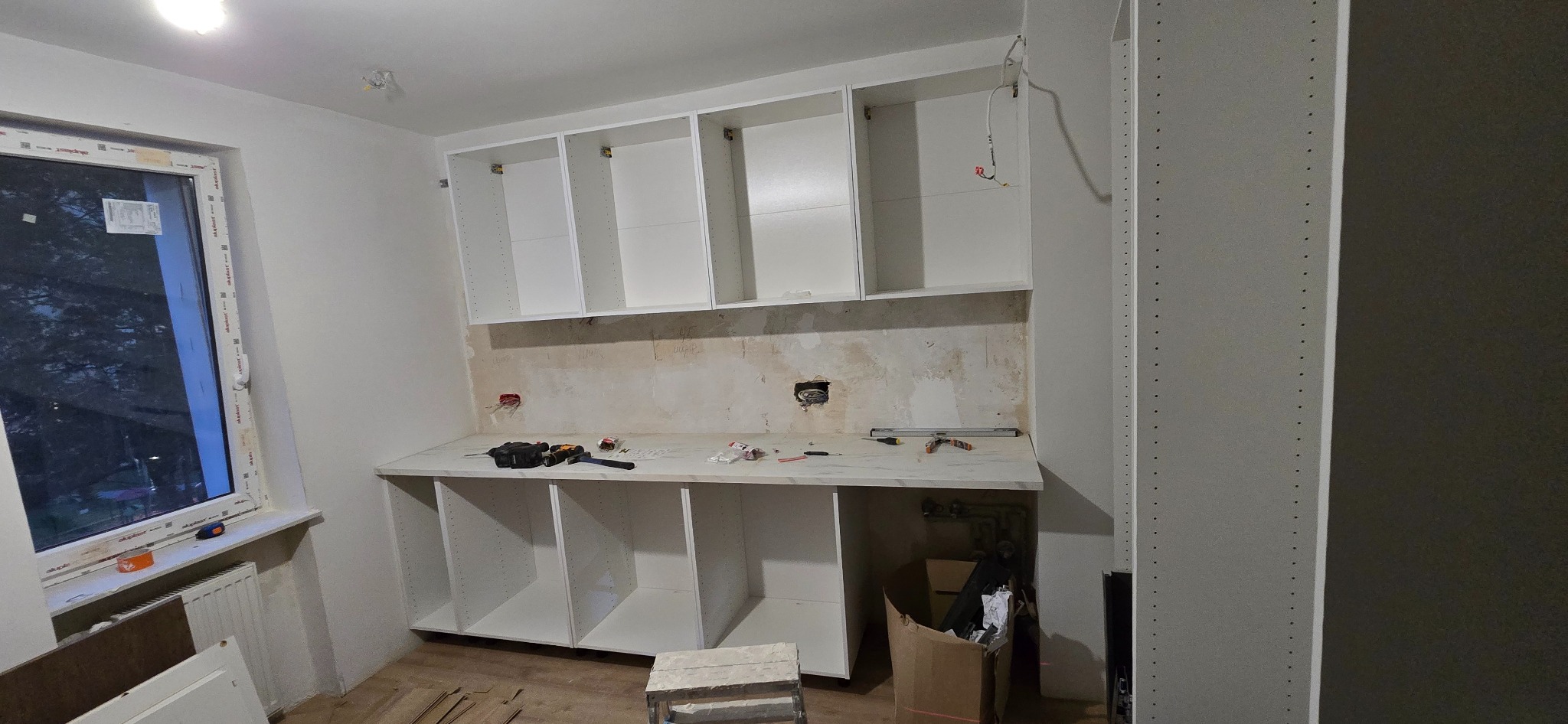 Montaż białych szafek kuchennych na ścianie, blat z narzędziami. Widoczne puszki elektryczne i pozostałości po remoncie. W tle okno. Widok na kuchnię w trakcie montażu.