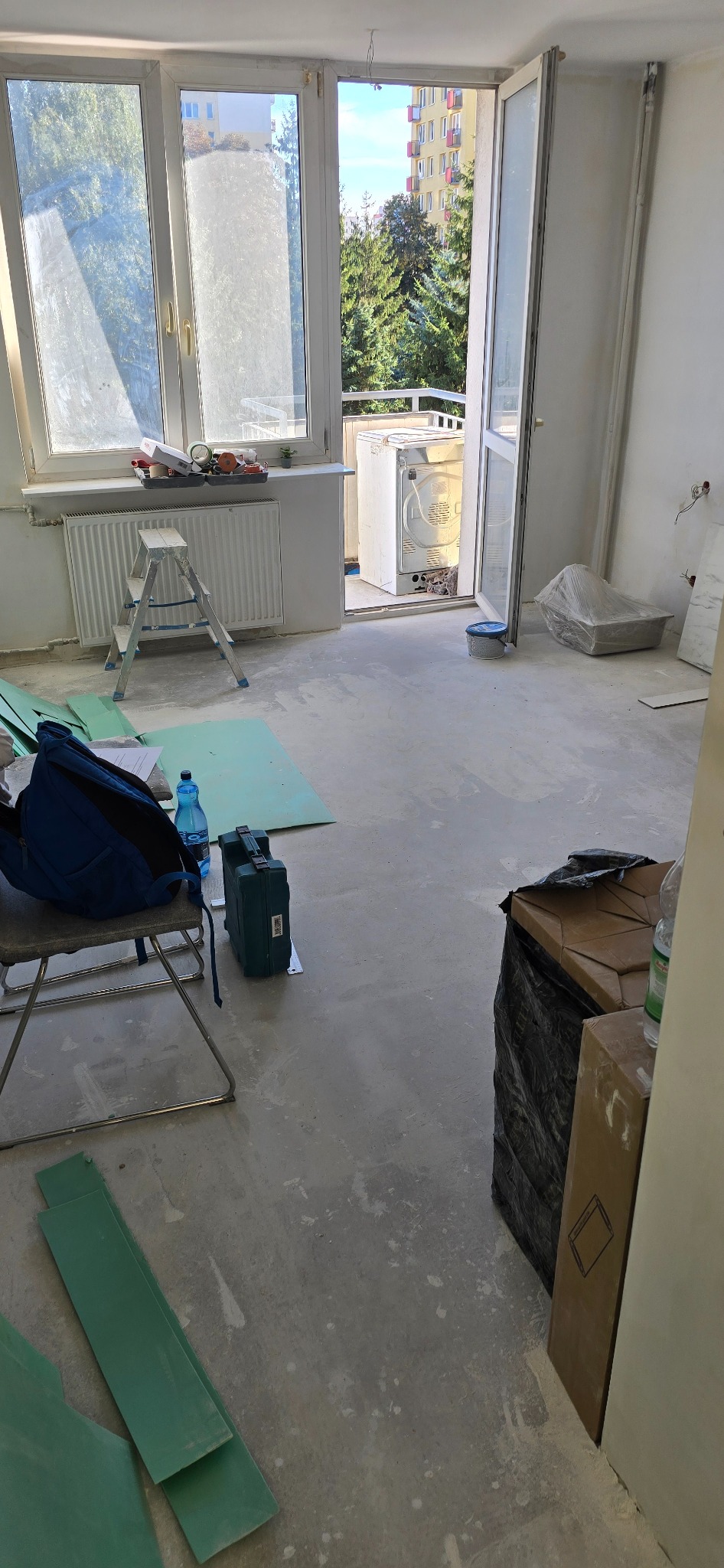 Wnętrze w trakcie remontu: drabina, narzędzia, materiały podłogowe, otwarte drzwi balkonowe z widokiem na zieleń i budynek. Podłoga przygotowana pod układanie.