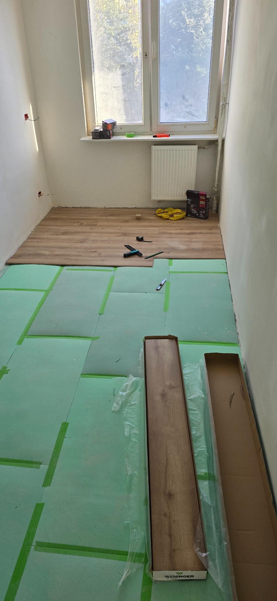 Układanie paneli podłogowych w pokoju: zielona podkładka, częściowo ułożone panele, narzędzia i otwarte kartony z panelami przy ścianie z oknem.