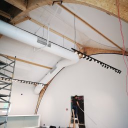 Energo-instal - Montaż oświetlenia szynowego w wysokim pomieszczeniu z drewnianą konstrukcją stropu. Elektryk na drabinie instaluje lampy na tle białej ściany.