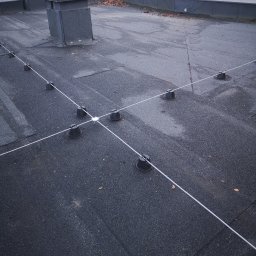 Energo-instal - Szczegółowe ujęcie dachu z zamontowaną instalacją odgromową, widoczne czarne uchwyty i białe przewody tworzące siatkę. Dach pokryty papą, z kominem w tle.