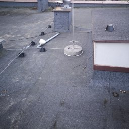 Energo-instal - Instalacja odgromowa na dachu pokrytym papą: maszt z uziomem, przewody i uchwyty. Widoczny komin i świetlik dachowy. Szare tło dachu z drobnymi zanieczyszczeniami.