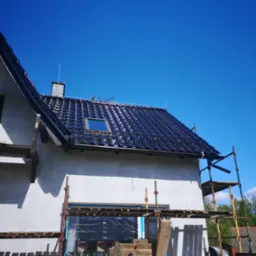 Dom w trakcie budowy z czarną dachówką, widoczne rusztowania i niezabezpieczone okno. Na dachu zamontowany maszt odgromowy.