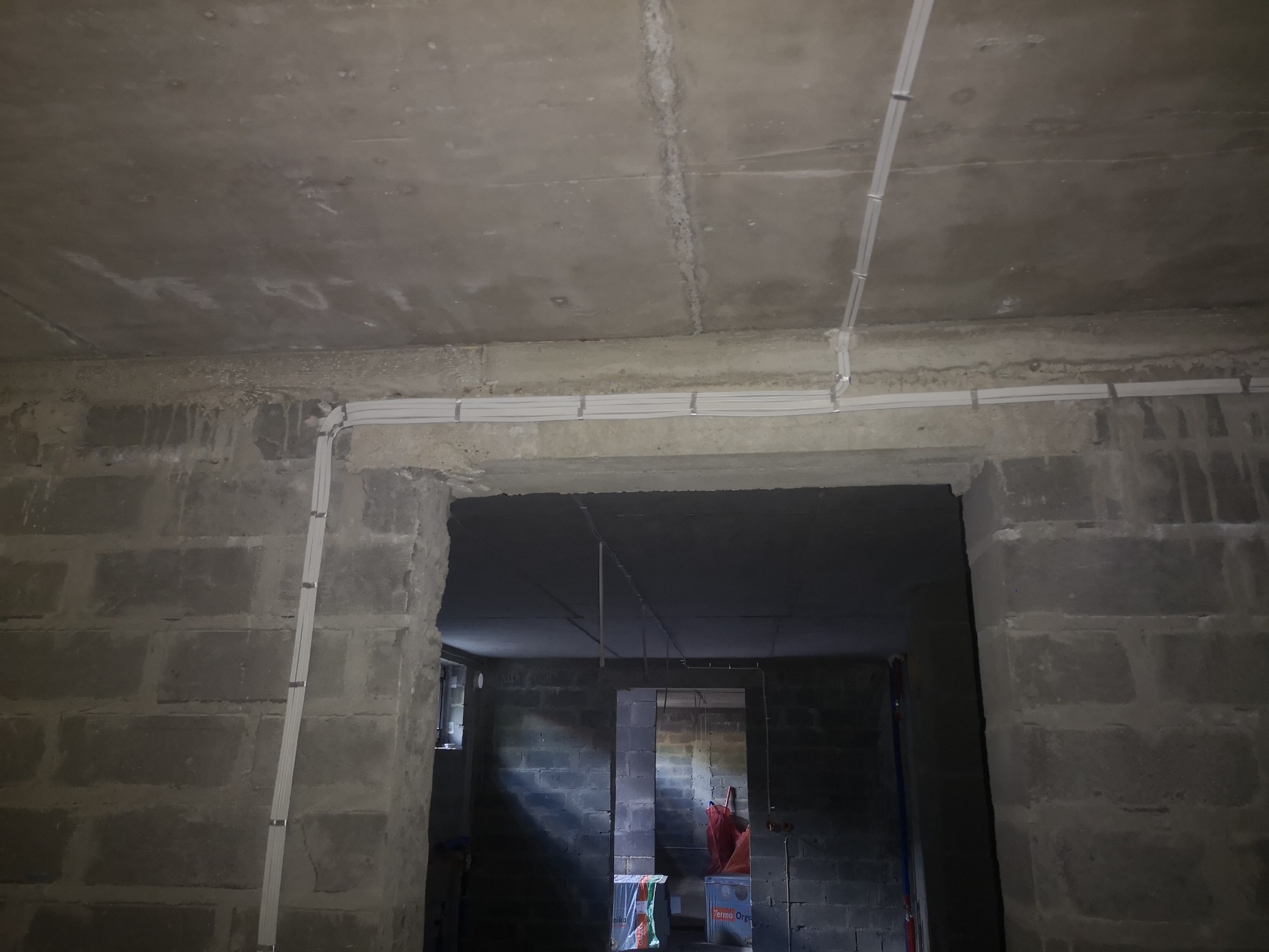 Szare ściany z betonu komórkowego z widocznymi przewodami elektrycznymi, zamocowanymi do sufitu i ścian. W tle widoczne kolejne pomieszczenie w stanie surowym.
