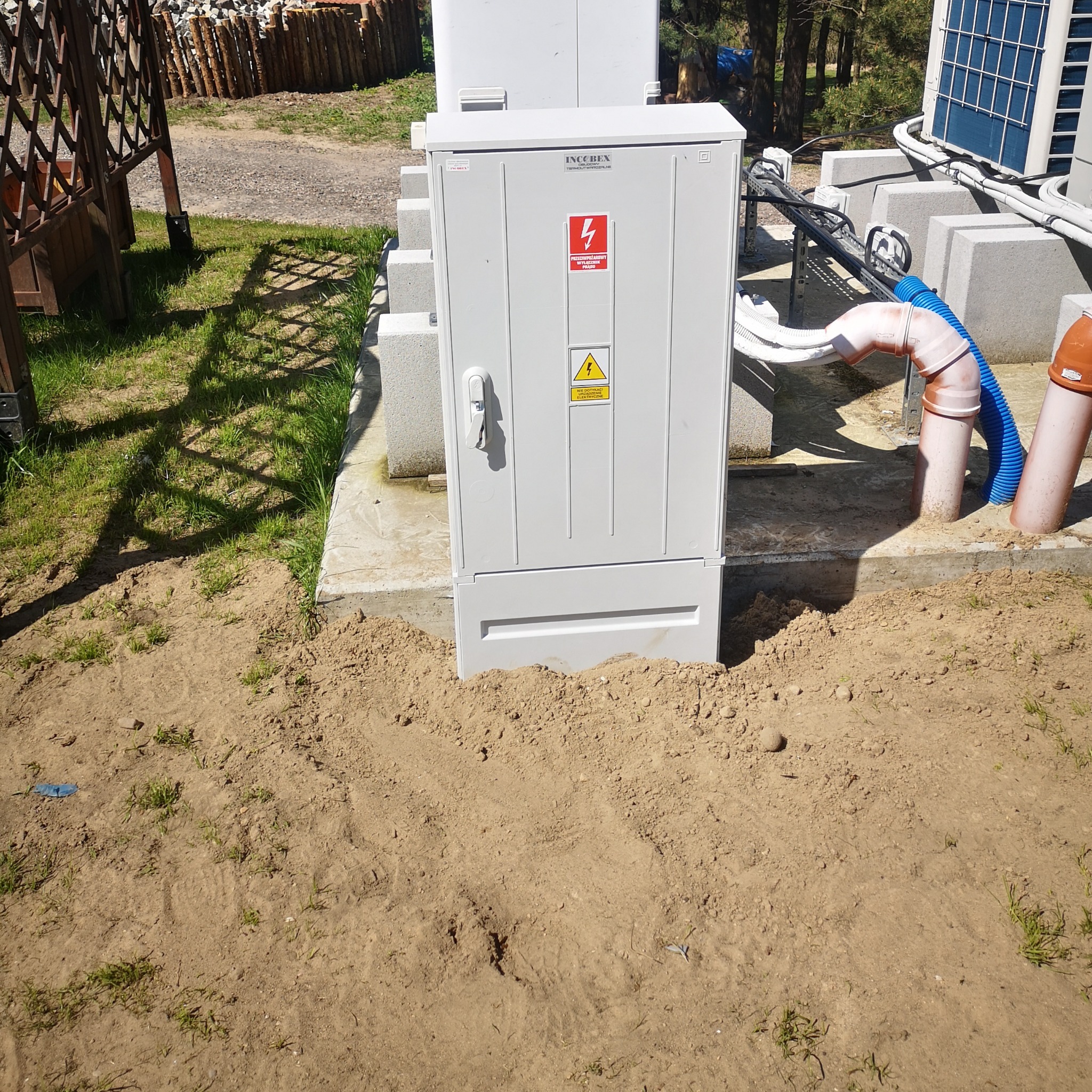 Szara szafa elektryczna z ostrzeżeniami, zamontowana na zewnątrz, otoczona ziemią. Widoczne kable i rury. Słońce oświetla kadr.