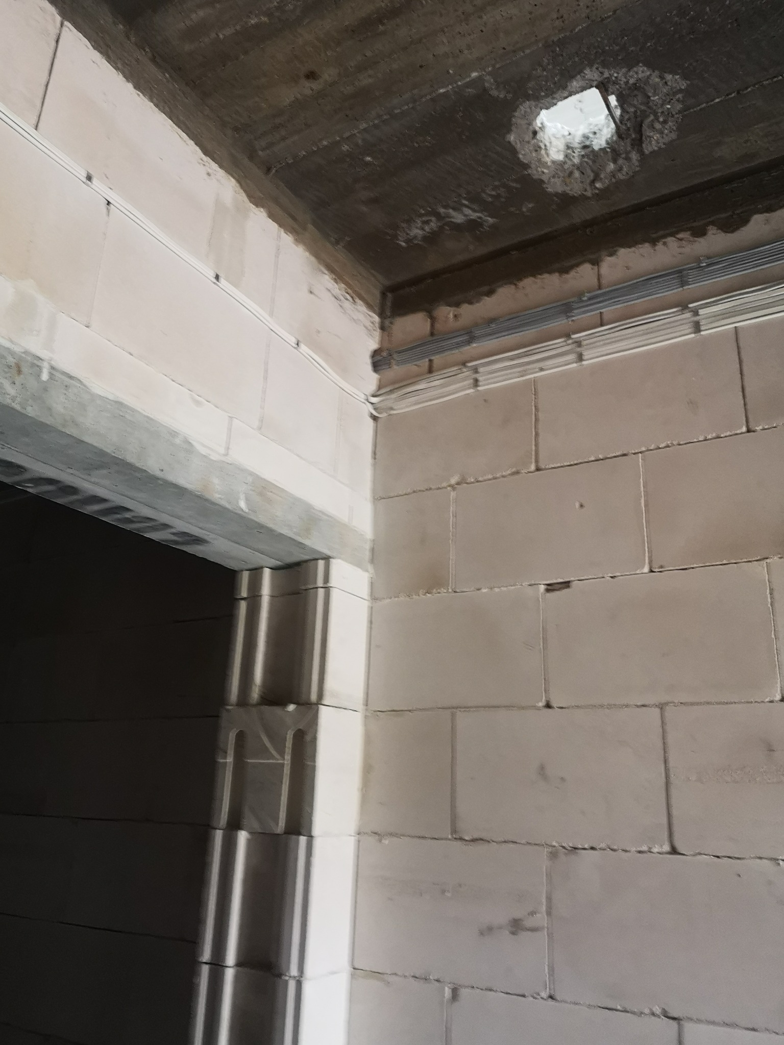 Szare rury elektryczne prowadzone po ścianie z białych bloczków, surowy betonowy sufit z otworem, widok z narożnika pomieszczenia w trakcie budowy.