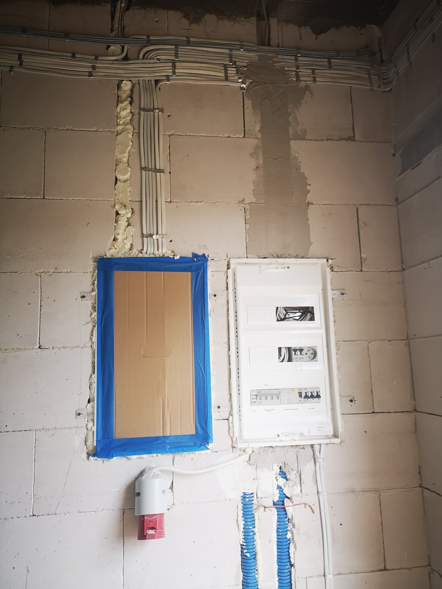 Biała ściana z betonu komórkowego z zamontowaną skrzynką elektryczną i zabezpieczonym otworem. Widoczne kable i rury instalacyjne w pianie montażowej.