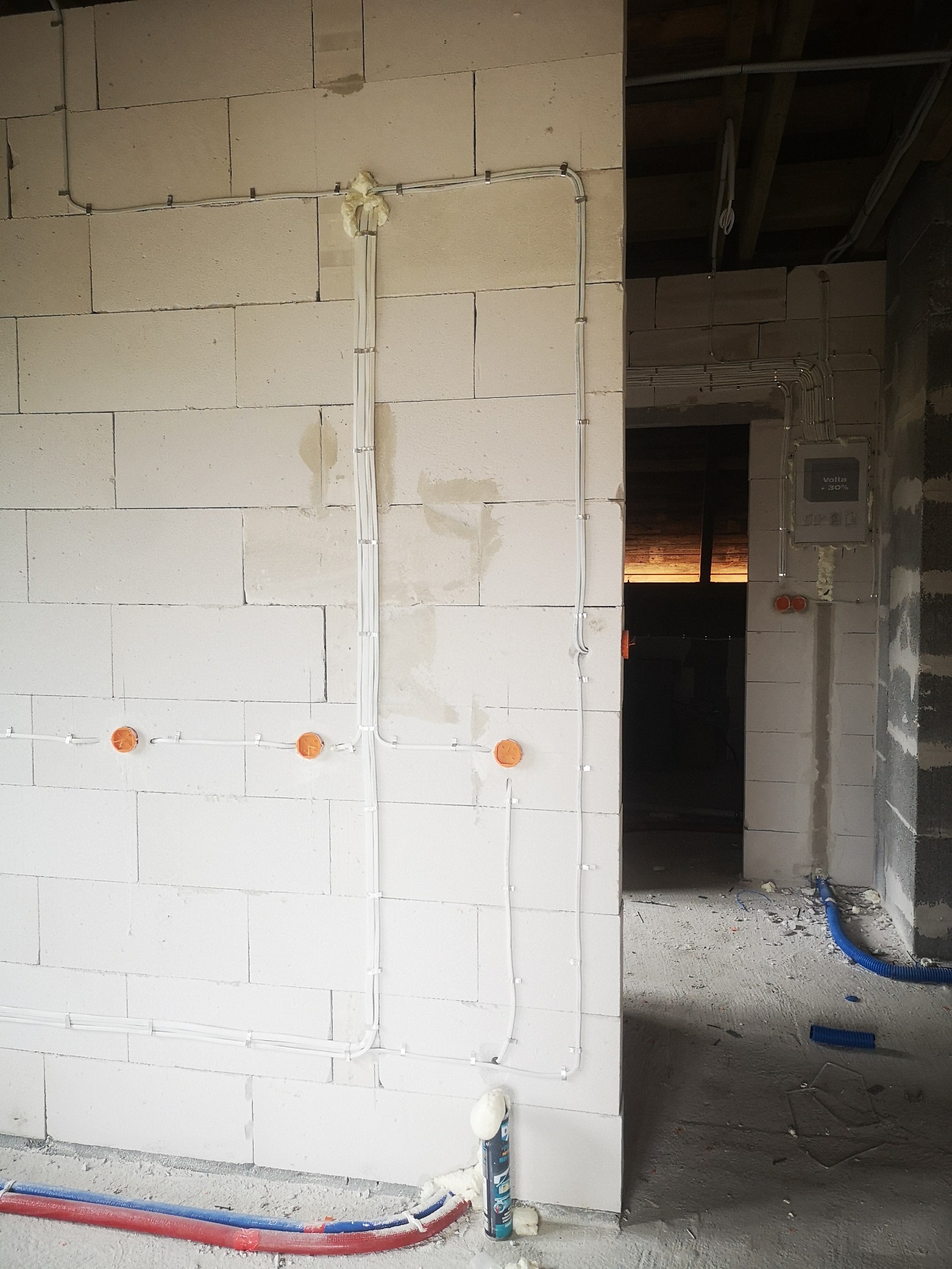 Białe przewody elektryczne przymocowane do ściany z białych bloczków, pomarańczowe puszki pod gniazdka, niebieskie rury w podłodze, stan surowy.