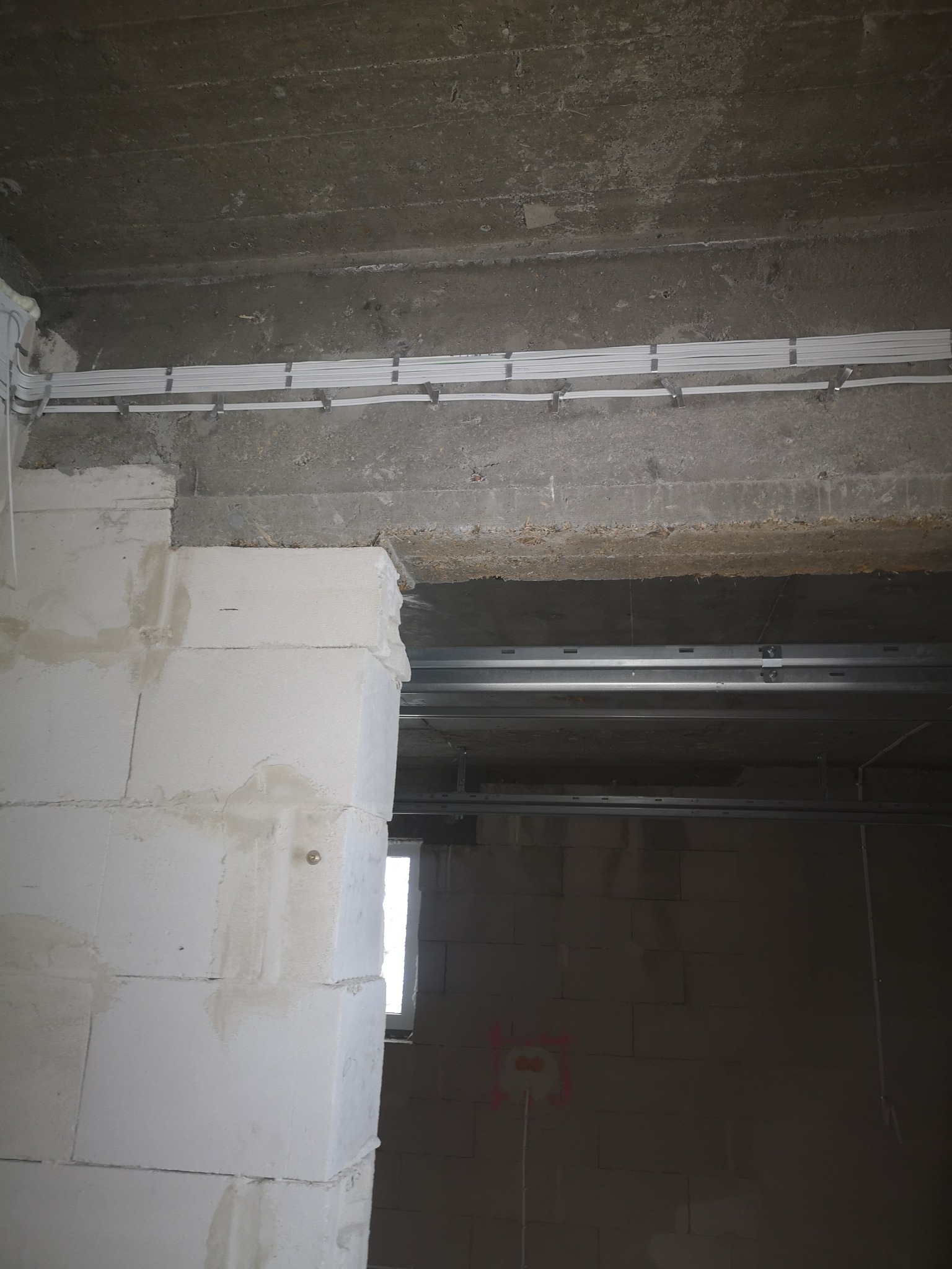 Białe kable elektryczne ułożone w korytkach na betonowym suficie w surowym wnętrzu. Widoczny fragment ściany z białych bloczków i metalowe profile podwieszanego sufitu.