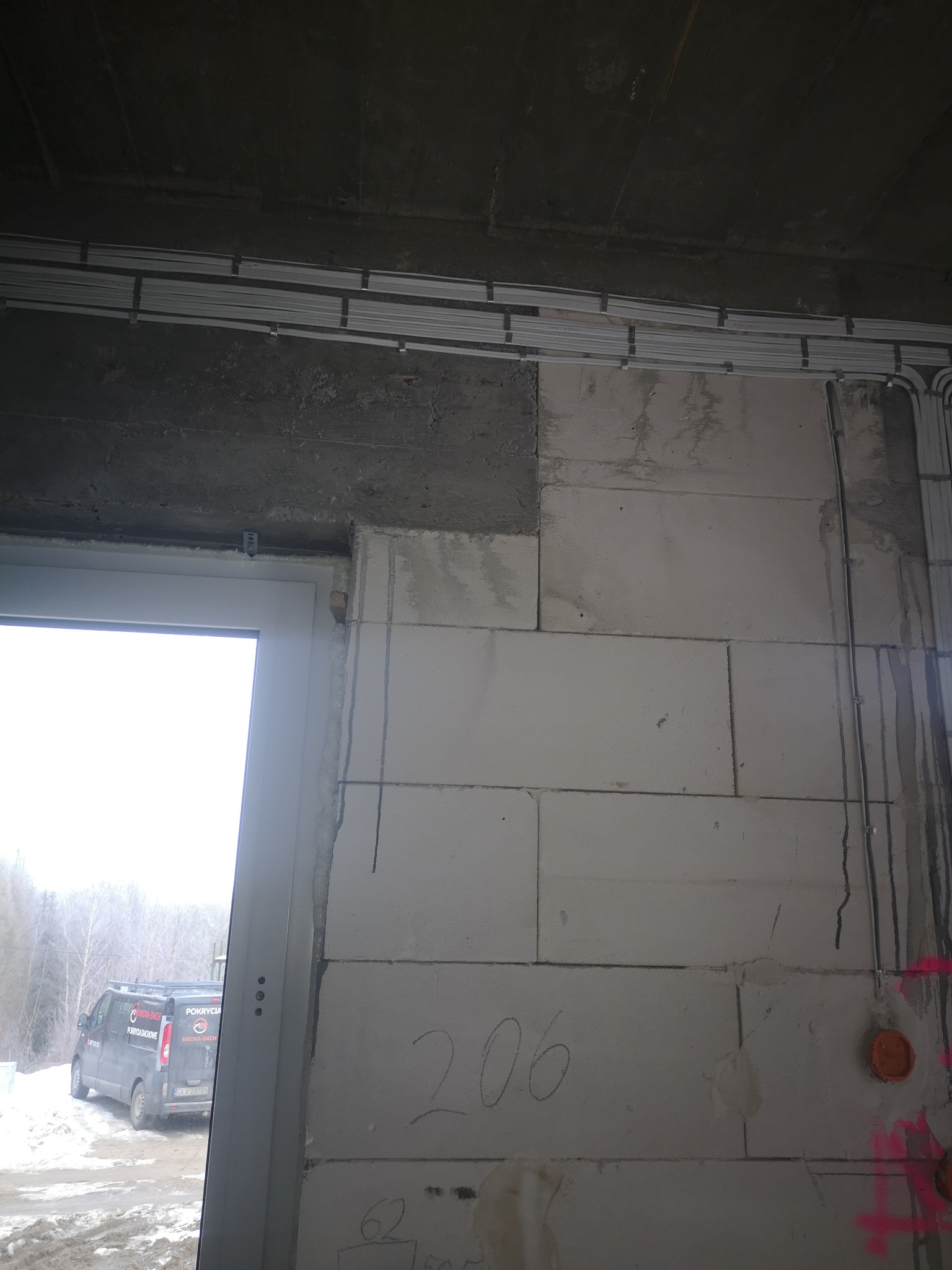 Wnętrze w stanie surowym: widoczne kable w korytkach na suficie, instalacja elektryczna na ścianie z bloczków, oznaczenia numerów pomieszczeń. Na zewnątrz widoczny samochód.