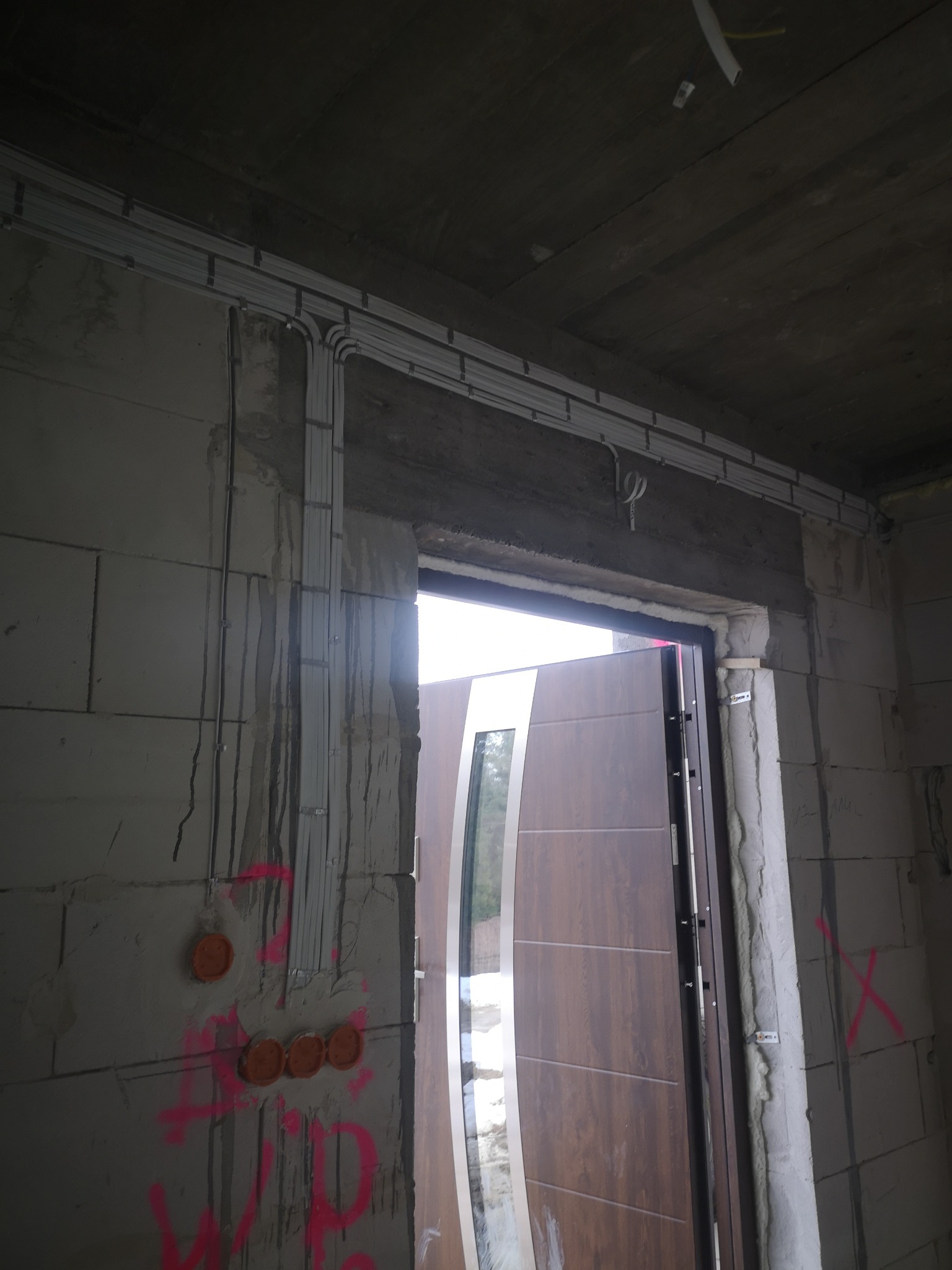 Surowy stan: Instalacja elektryczna w korytach kablowych nad futryną drzwiową, z widocznymi puszkami instalacyjnymi w ścianie z betonu komórkowego.