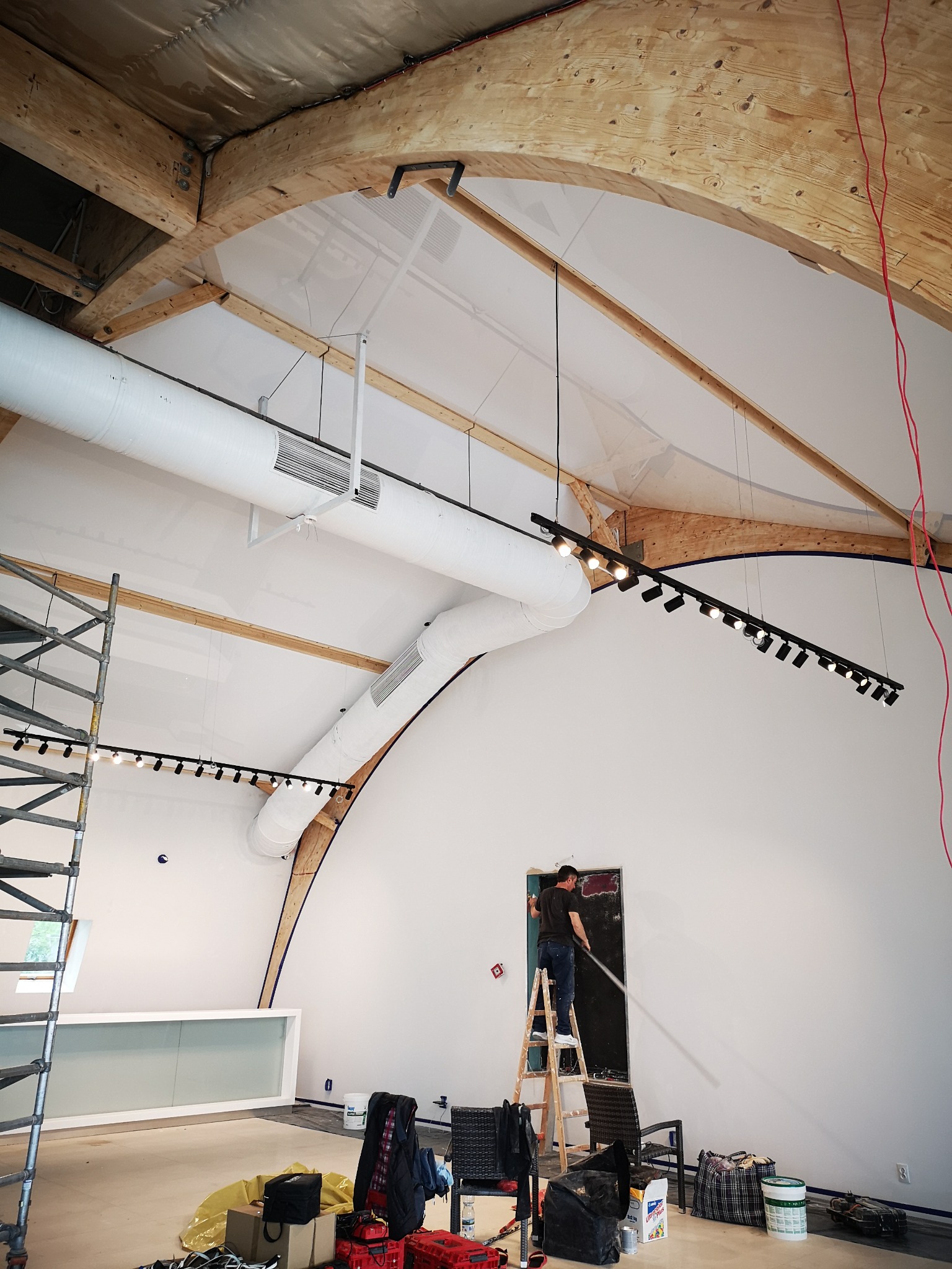 Montaż oświetlenia szynowego w wysokim pomieszczeniu z drewnianą konstrukcją stropu. Elektryk na drabinie instaluje lampy na tle białej ściany.