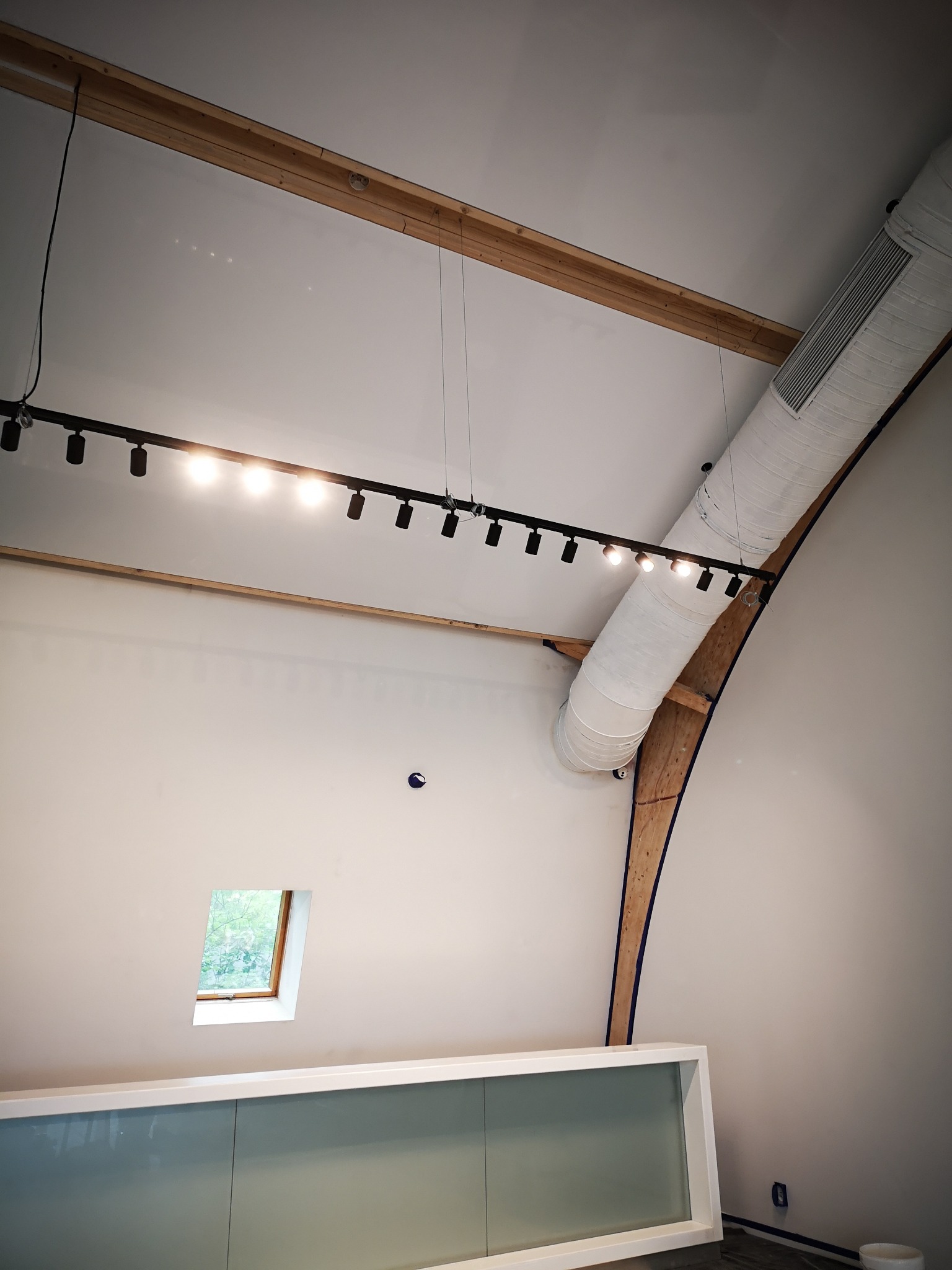 Wnętrze z oświetleniem szynowym i elementami instalacji wentylacyjnej. Widoczne drewniane elementy konstrukcyjne i niewielkie okno. Minimalistyczny design.