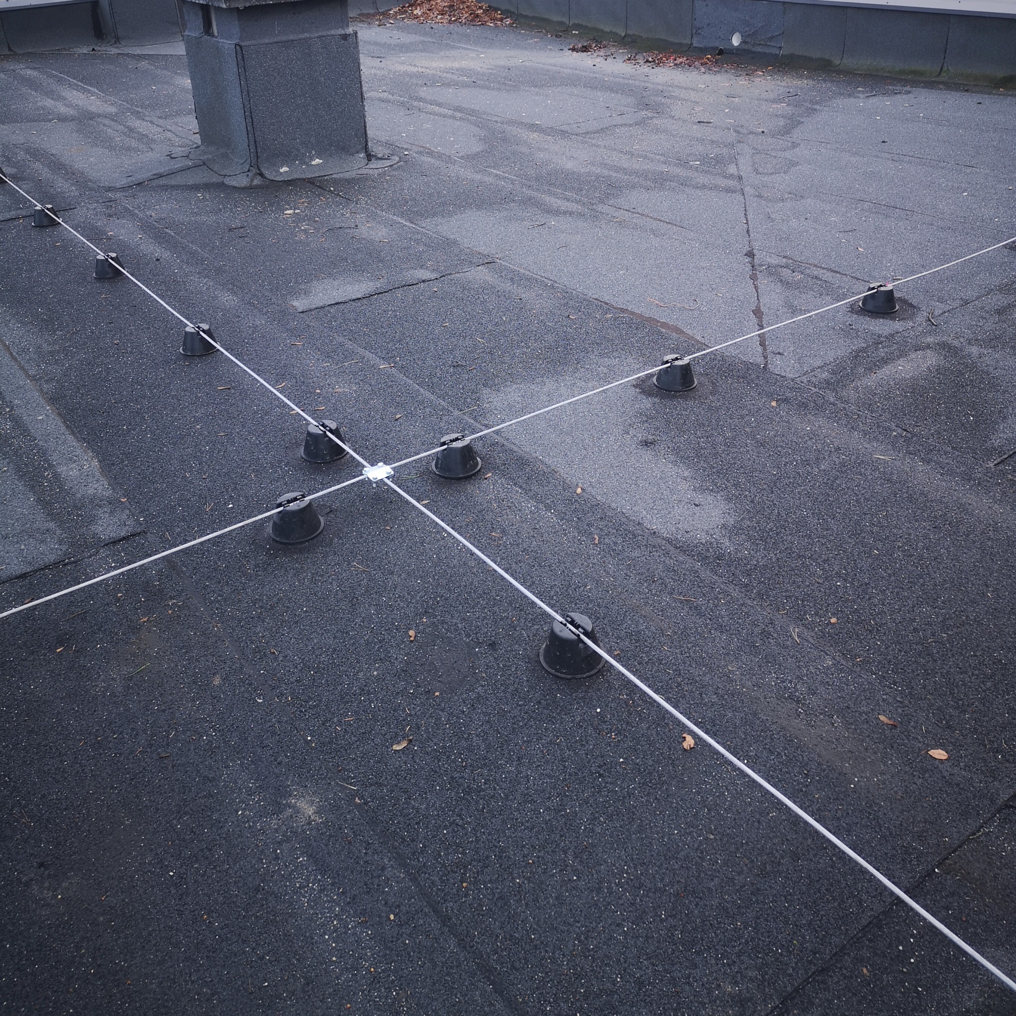 Szczegółowe ujęcie dachu z zamontowaną instalacją odgromową, widoczne czarne uchwyty i białe przewody tworzące siatkę. Dach pokryty papą, z kominem w tle.