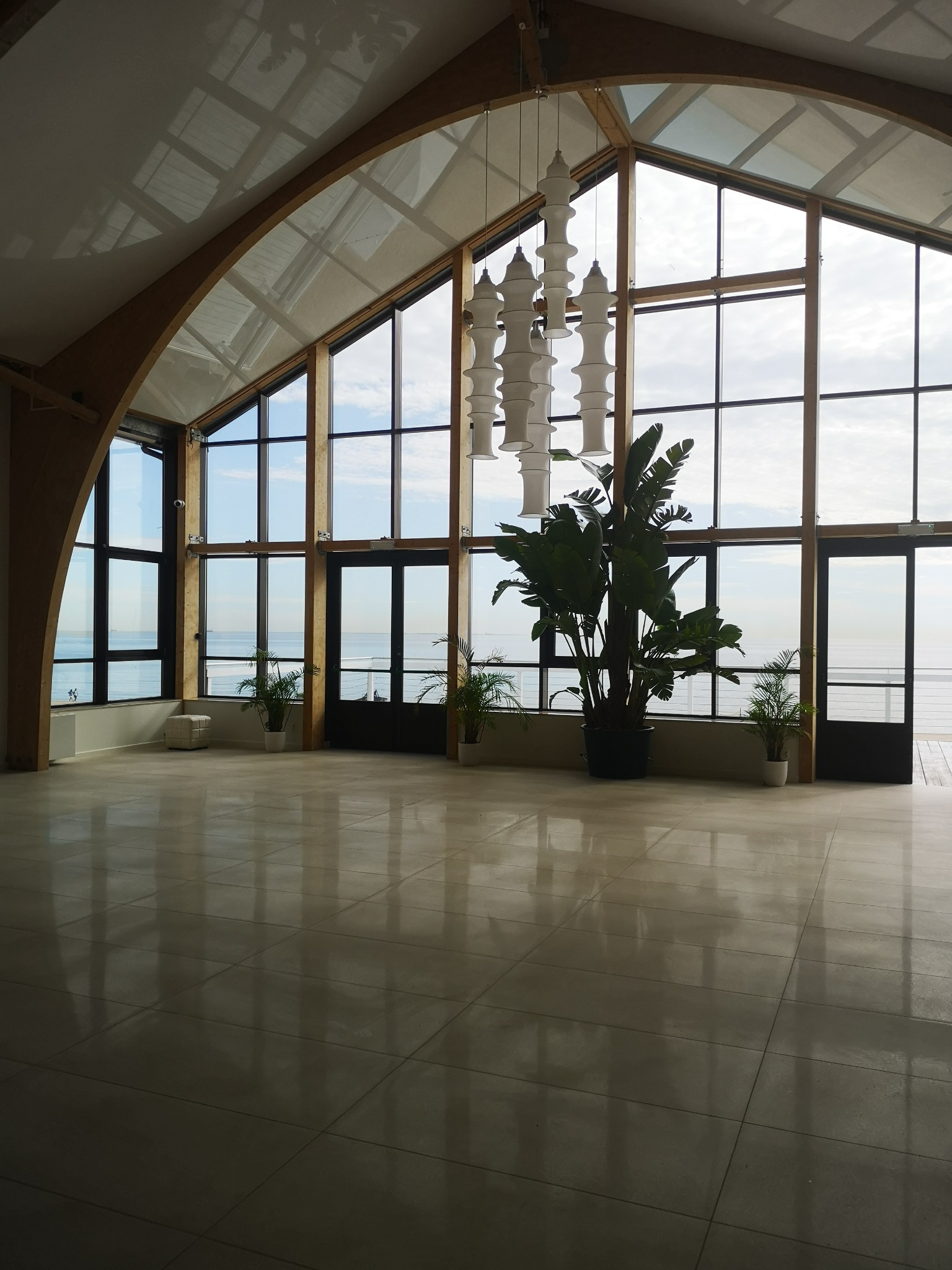 Przestronne wnętrze z wysokimi oknami wychodzącymi na morze. Widoczna nowoczesna lampa wisząca i rośliny doniczkowe. Jasna podłoga z płytek.