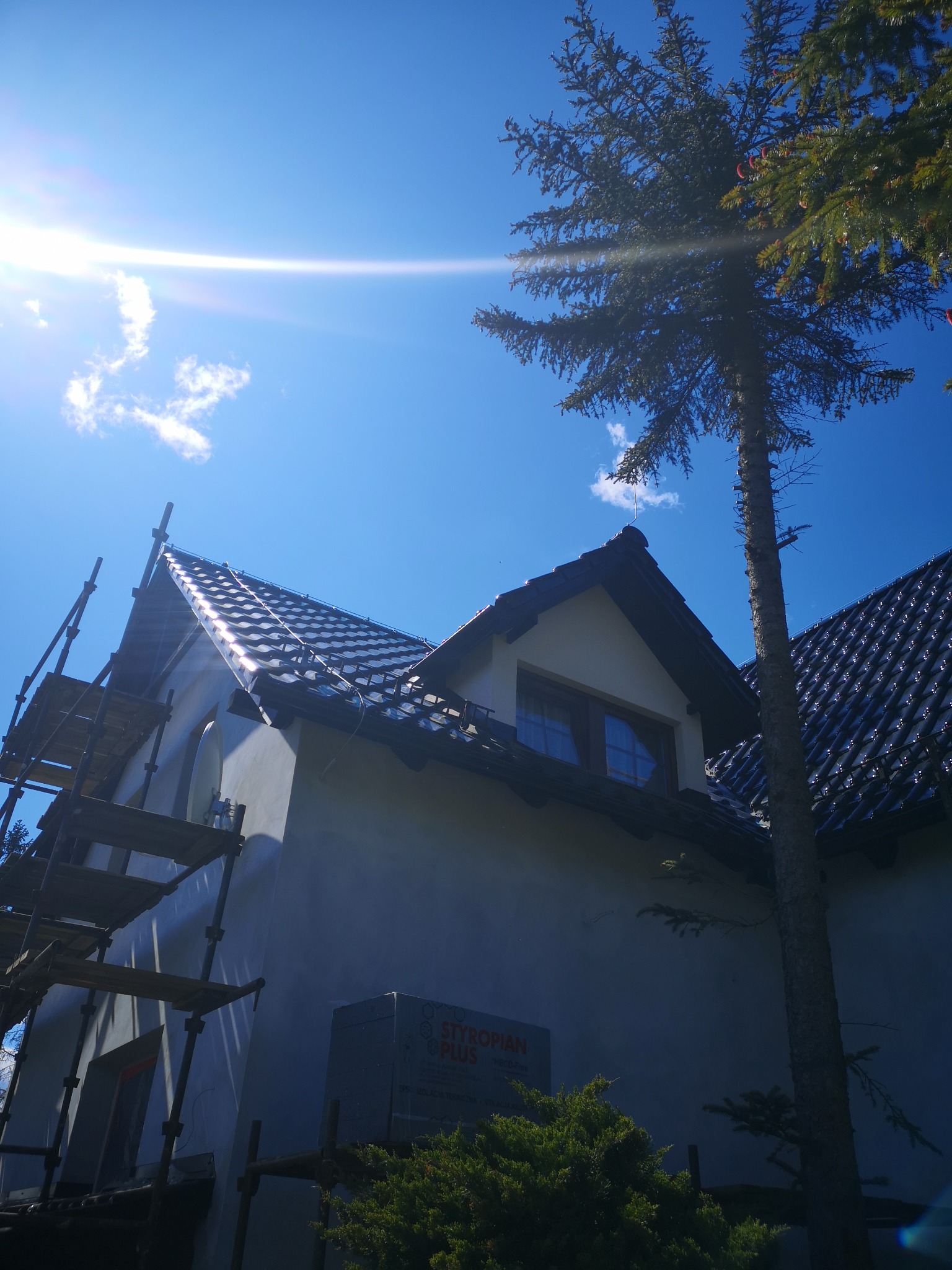 Dom w trakcie ocieplania, widoczne rusztowania i styropian. Błękitne niebo z promieniami słońca. Częściowo otynkowana fasada i ciemna dachówka.