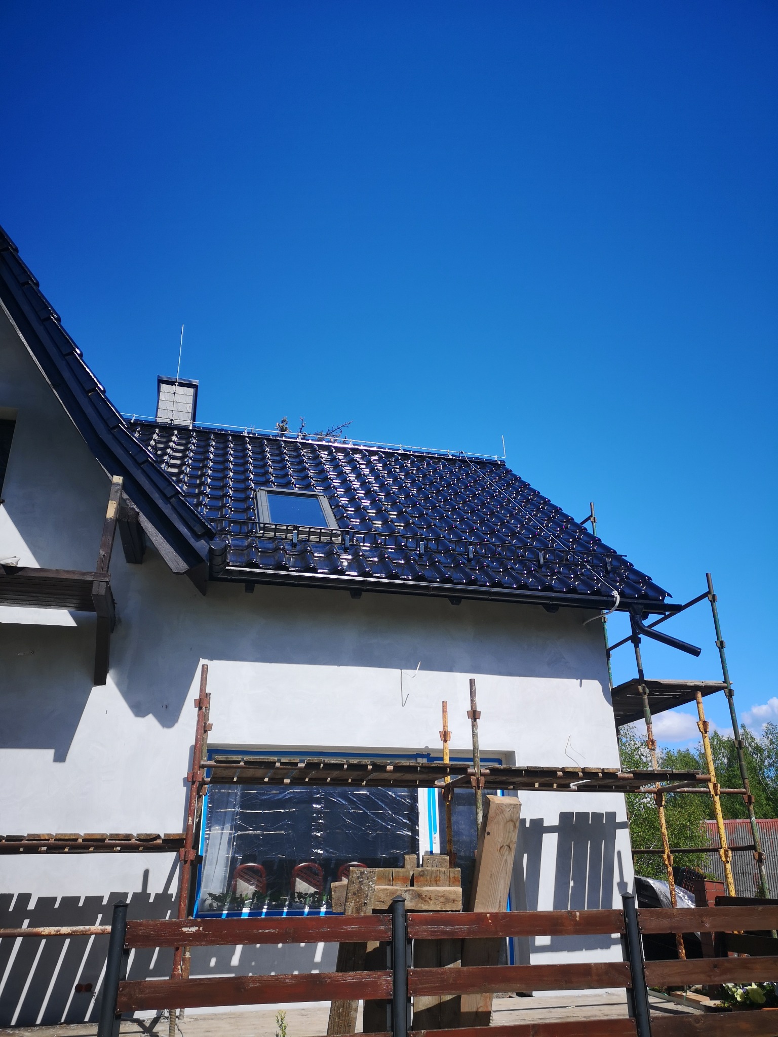 Dom w trakcie budowy z czarną dachówką, widoczne rusztowania i niezabezpieczone okno. Na dachu zamontowany maszt odgromowy.