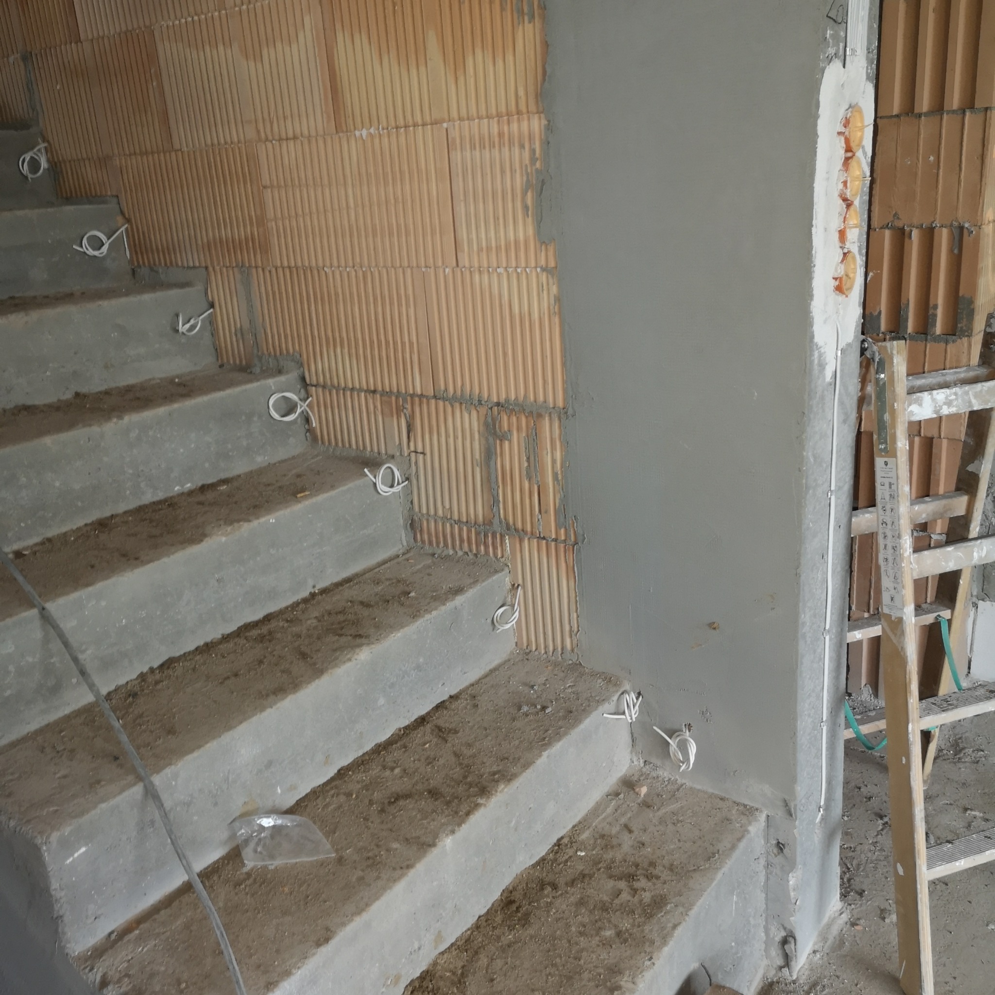 Surowe betonowe schody z przewodami elektrycznymi wystającymi ze ściany z pustaków, widoczne puszki instalacyjne i drabina. Etap prac instalacyjnych.