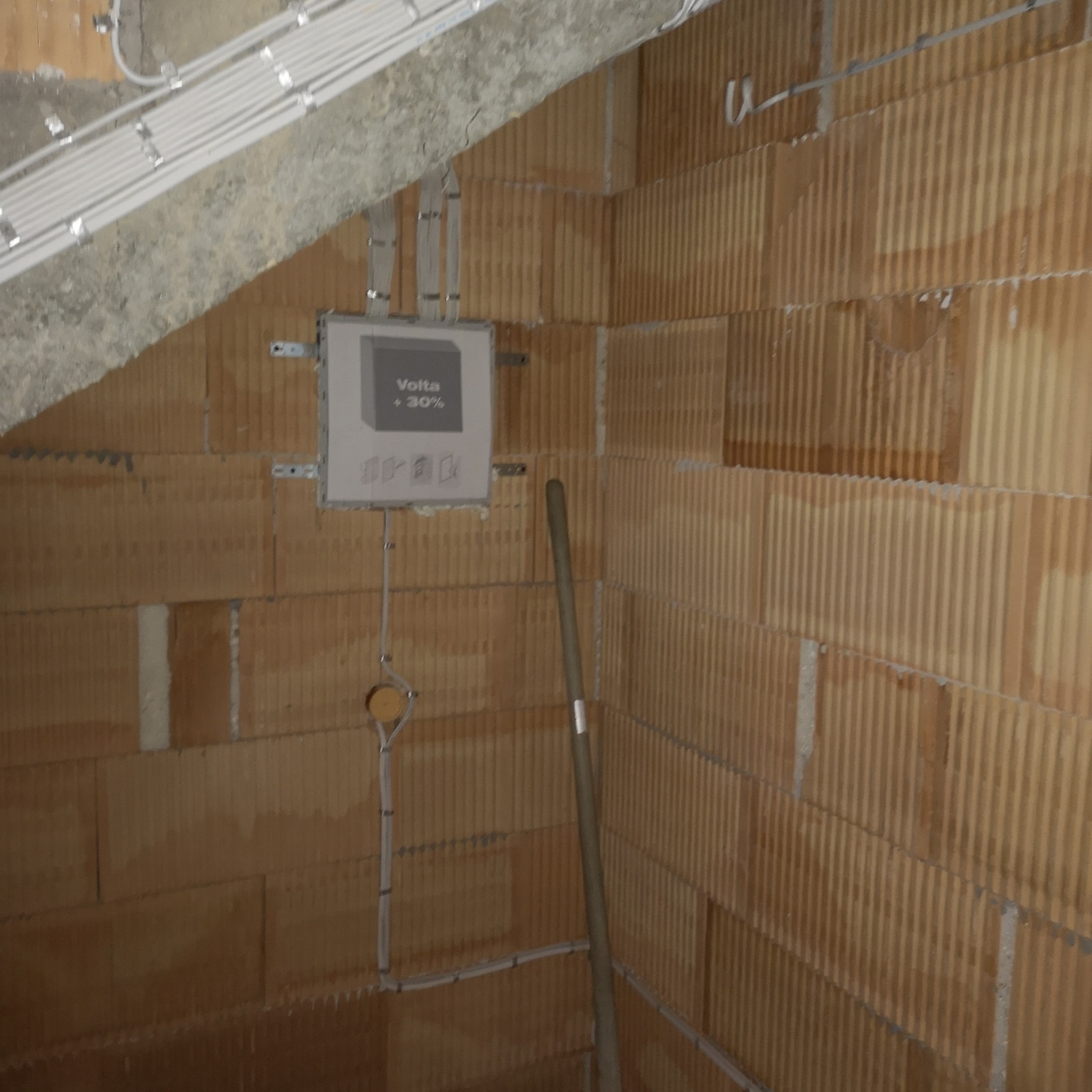 Rozdzielnica elektryczna Volta +30% zamontowana na ścianie z bloczków, z widocznymi przewodami i peszlami. Instalacja w trakcie prac, widoczny fragment ściany i sufitu.