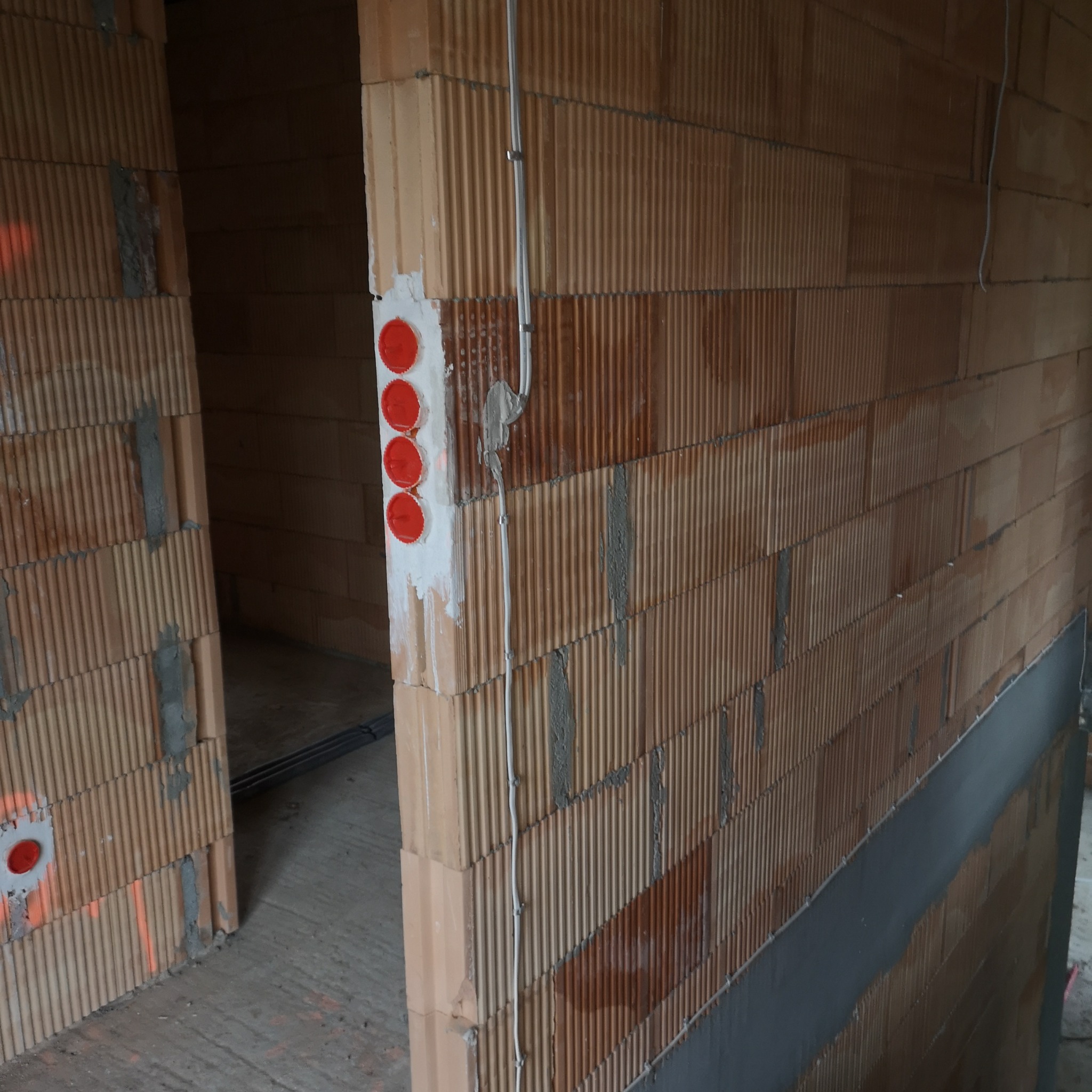 Ściana z cegły dziurawki z zamontowanymi przewodami elektrycznymi i puszkami pod gniazdka z czerwonymi zaślepkami, widok na surowy stan budynku.