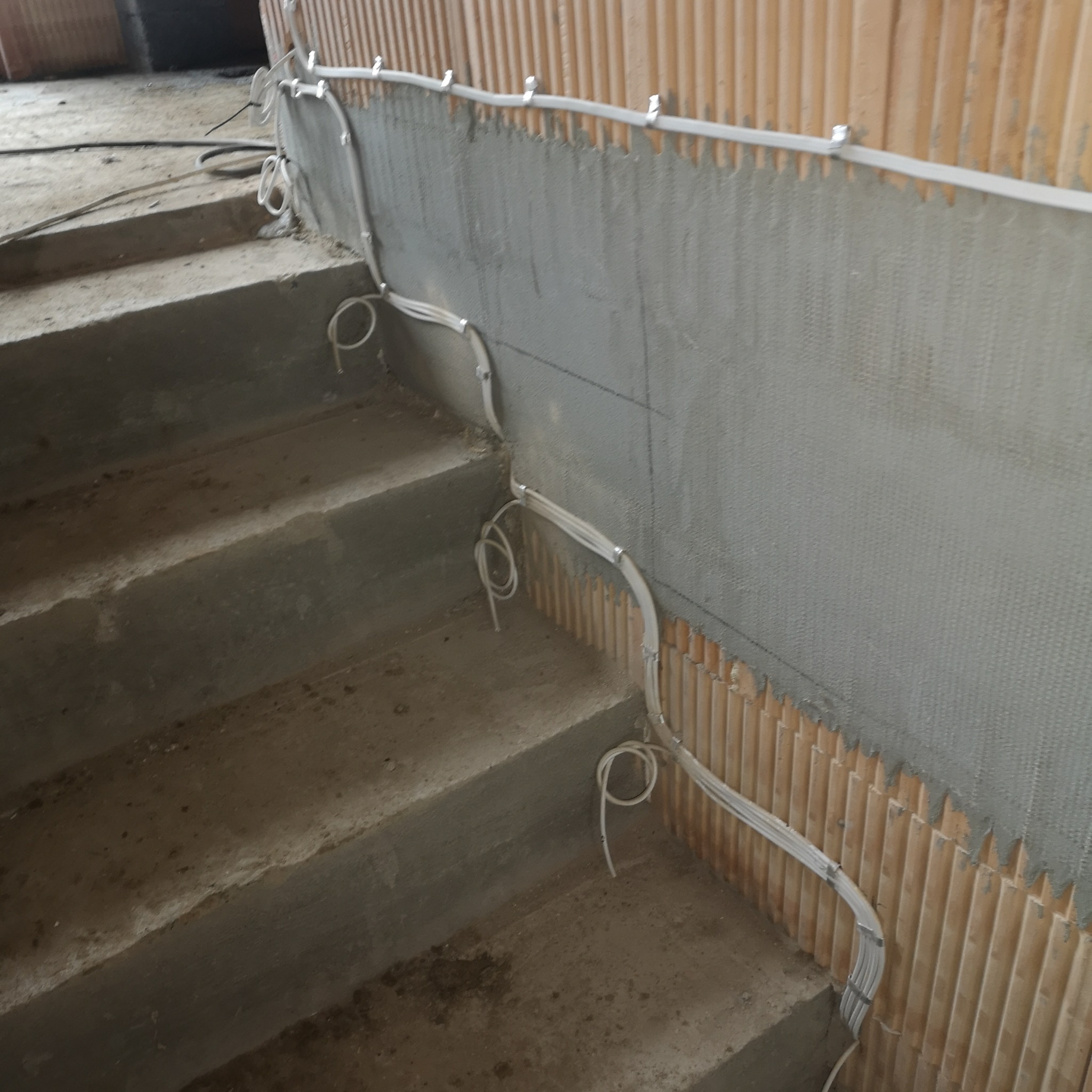 Instalacja kabli elektrycznych w korycie na ścianie przy schodach. Kable przymocowane klipsami. Surowy stan wykończenia ścian i schodów.