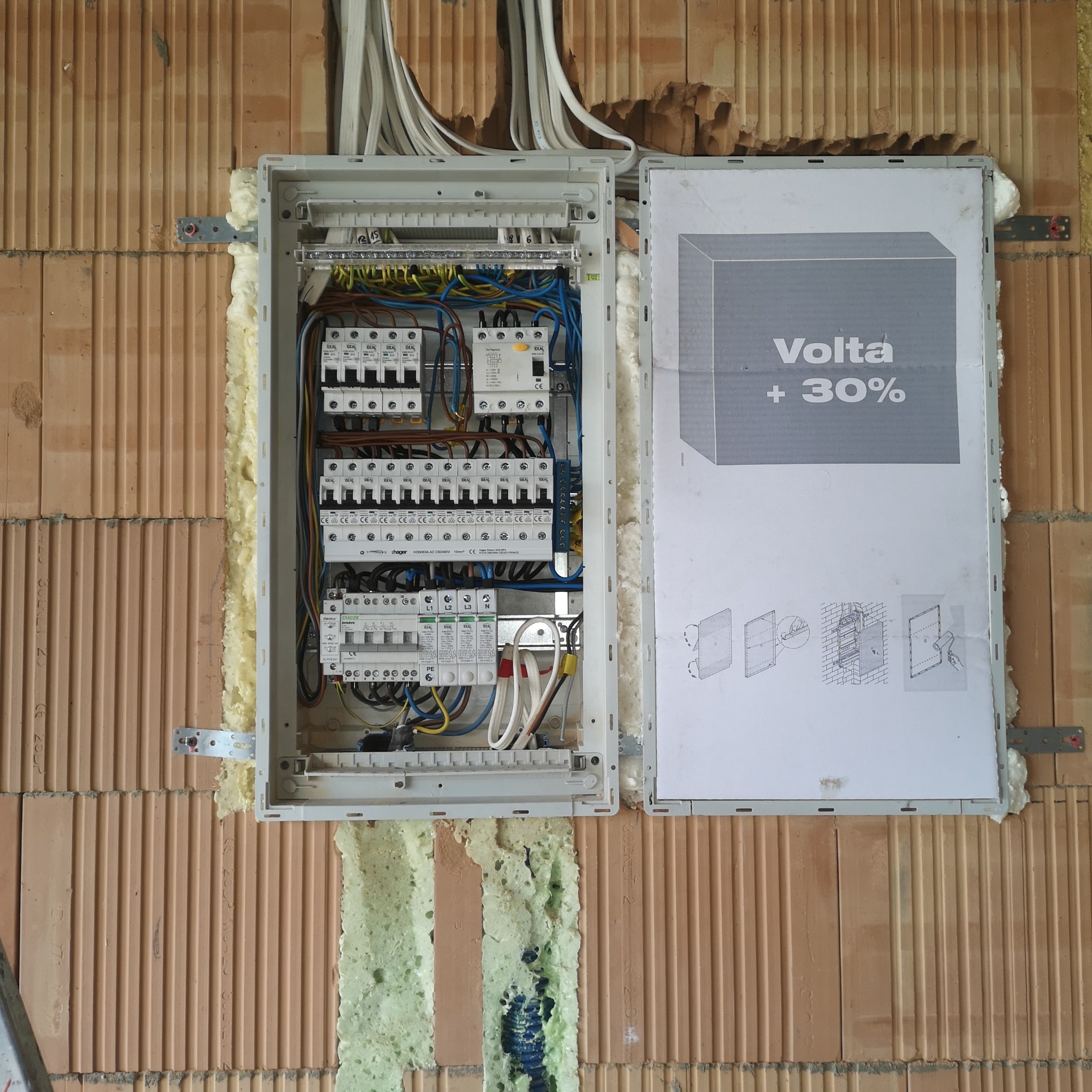 Otwarta skrzynka elektryczna z bezpiecznikami i przewodami, zamontowana na murowanej ścianie z cegły, uszczelniona pianką montażową, z widoczną instrukcją Volta +30%.
