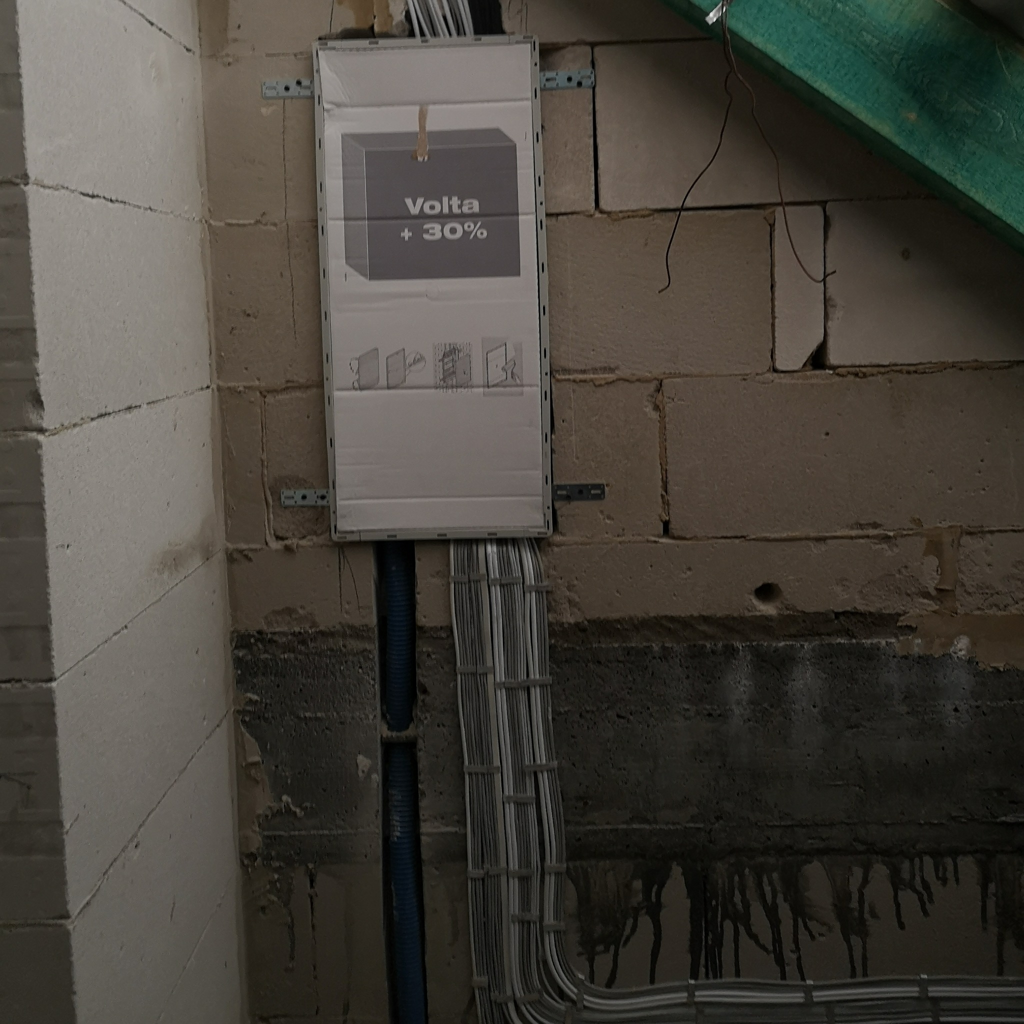 Skrzynka elektryczna Volta +30% zamontowana na ścianie z bloczków, z widocznymi przewodami w peszlach i korytkach. Instalacja w surowym stanie.