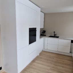 Michal Zolopa - Minimalistyczna, biała kuchnia z zabudową po sufit i czarnymi akcentami armatury. Jasna podłoga z paneli imitujących drewno. Widok na zlewozmywak i ekspres do kawy.