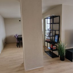 Michal Zolopa - Jasne wnętrze po odświeżeniu: beżowe ściany, drewniana podłoga, czarne meble RTV i regał. W tle widoczny stół z fioletowymi krzesłami.