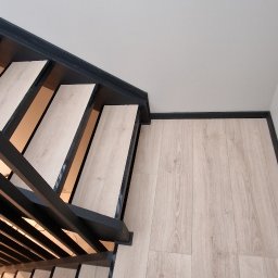 Michal Zolopa - Nowoczesne schody z drewnianymi stopniami i czarną metalową balustradą, dopasowane do jasnej podłogi. Minimalistyczny design i precyzyjne wykończenie.