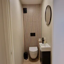 Wąska łazienka po remoncie: drewniane lamele na ścianie za toaletą, okrągłe lustro nad umywalką, podłoga wyłożona panelami. Nowoczesny design.